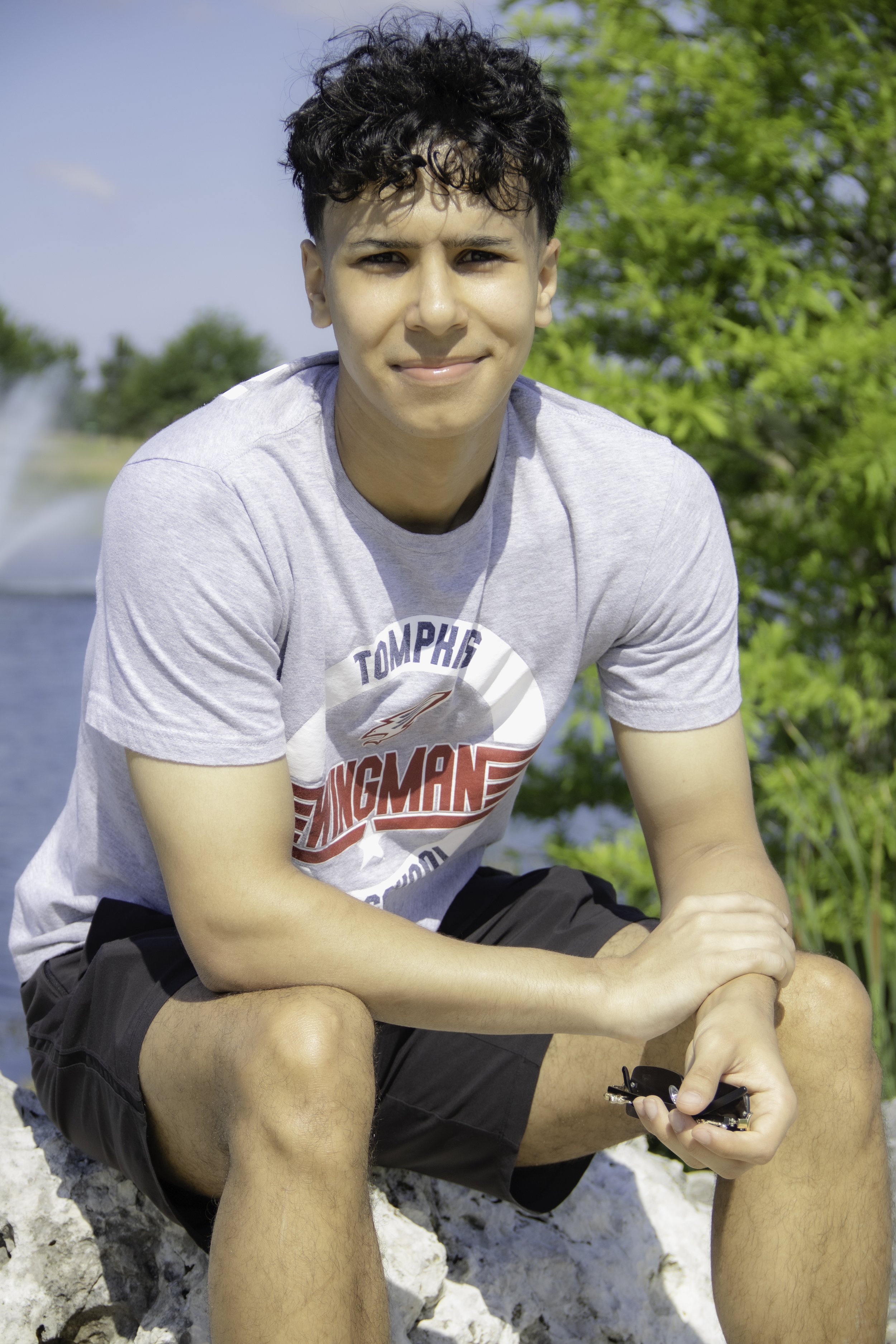 Young man sitting outdoors near water, wearing a gray T-shirt and black shorts, holding sunglasses, with green trees and blue sky in the background.
