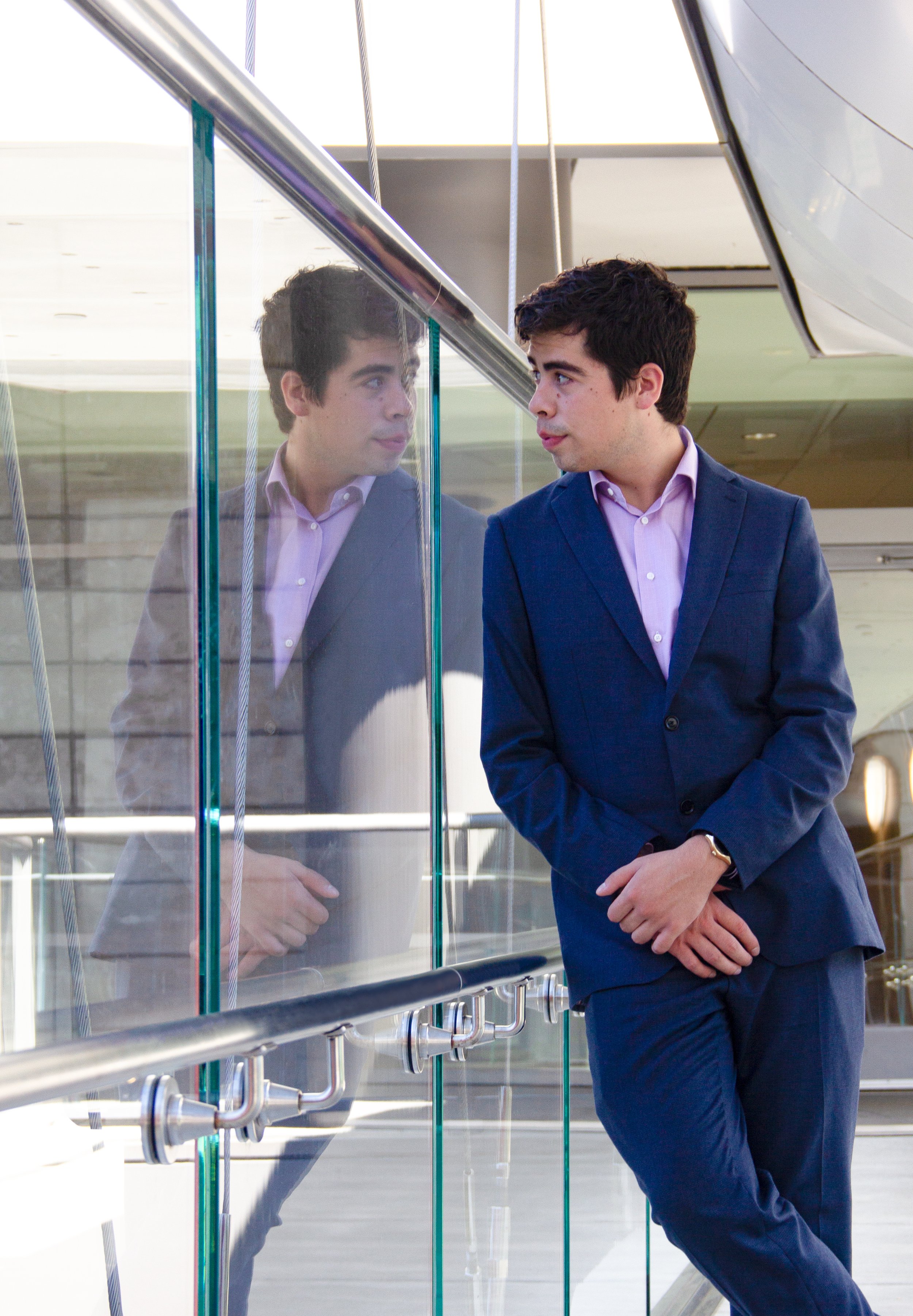 A man in a blue suit and pink shirt standing next to a glass barrier, looking at his reflection in the glass, in a modern indoor setting.