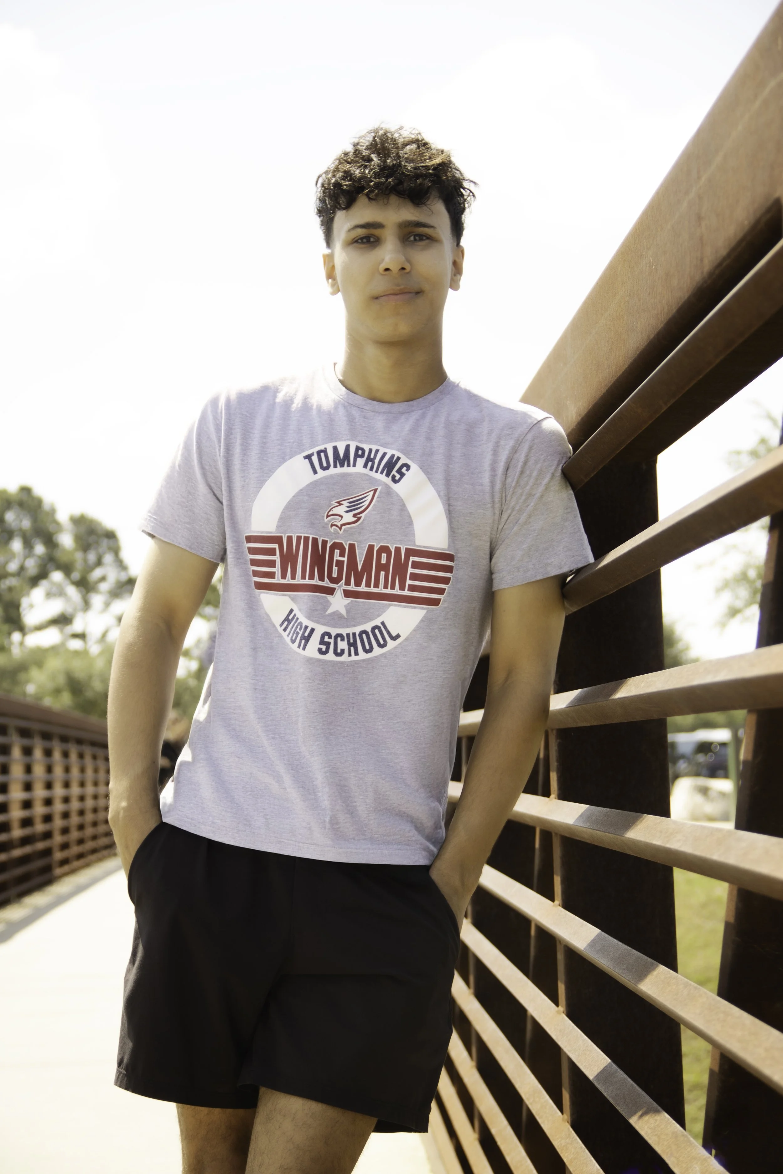 Young man with curly hair standing outdoors leaning against a wooden railing, wearing a gray T-shirt with 'Tompkins Wingman High School' logo and black shorts.