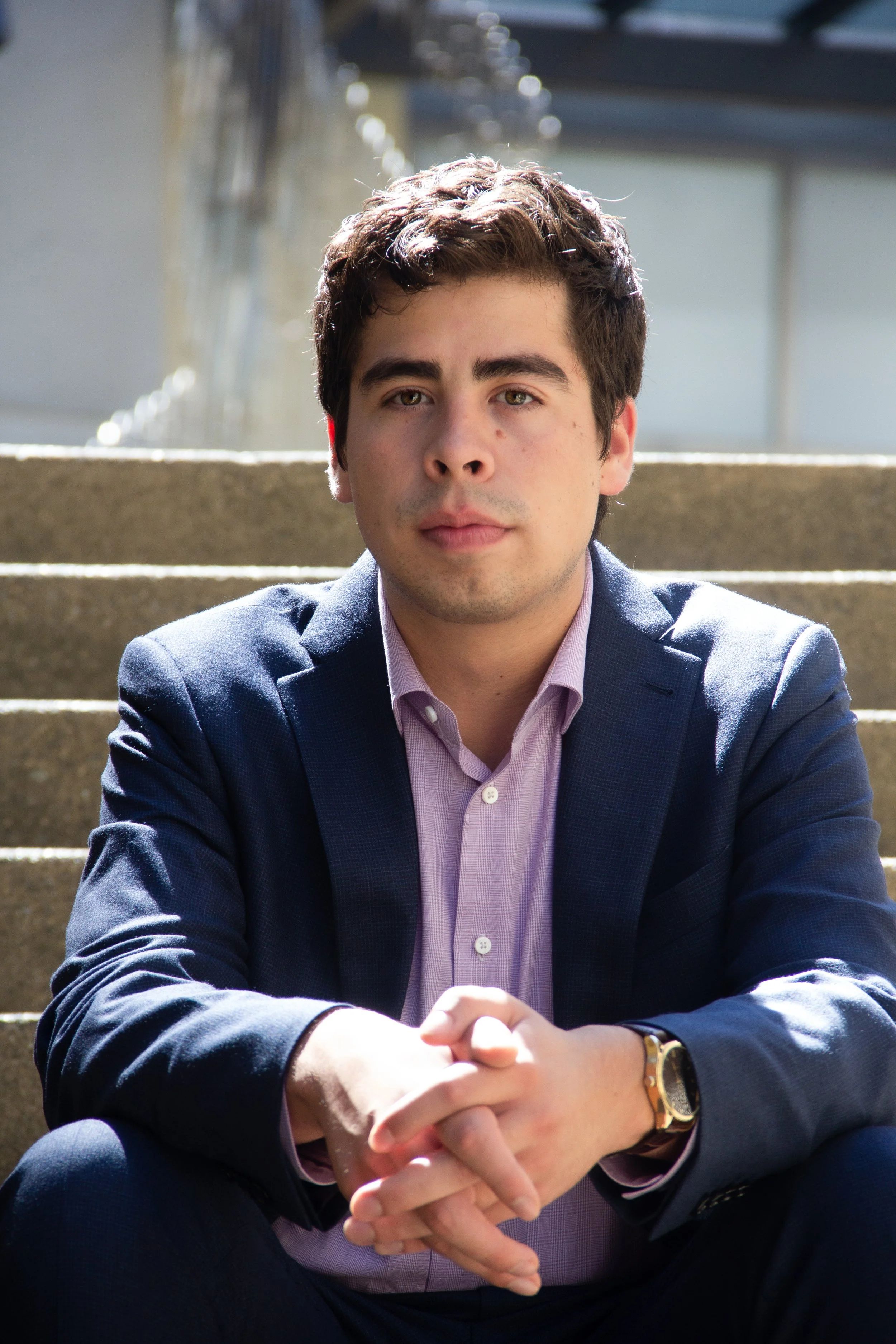 Young man in a navy blazer and pink shirt sitting on outdoor steps with hands clasped, looking directly at the camera.