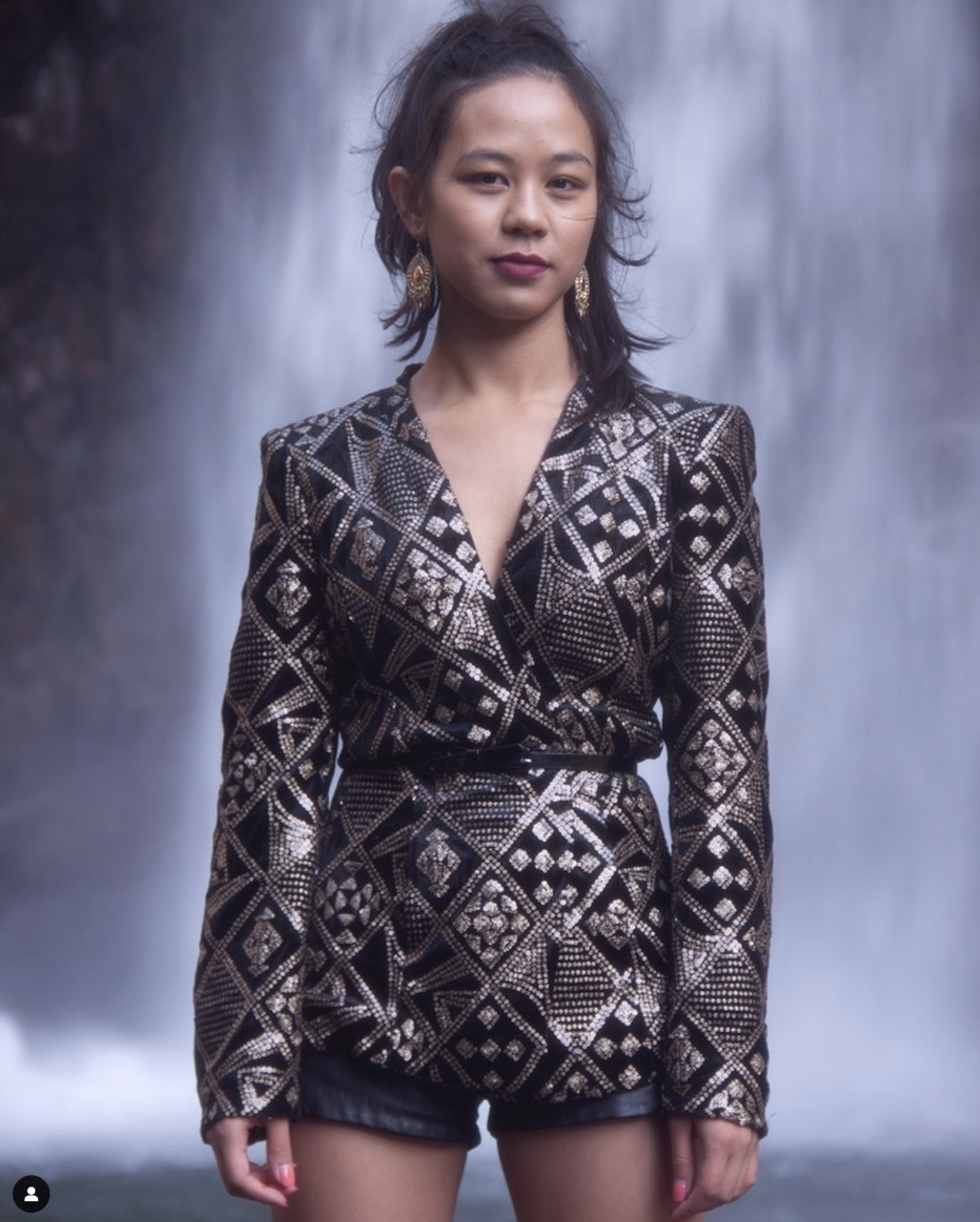 Person wearing a black and gold patterned outfit in front of a waterfall.