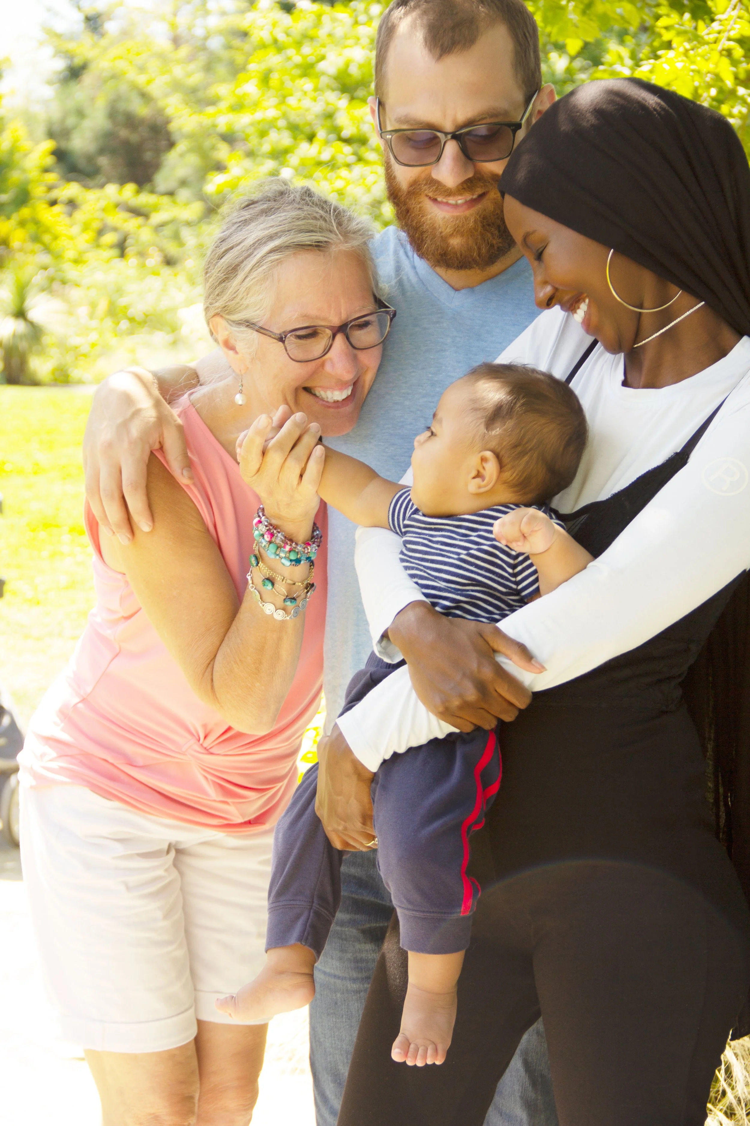 A multigenerational family, including an elderly woman, a young man, a woman with a hijab, and a baby, smiling and embracing each other outdoors on a sunny day.