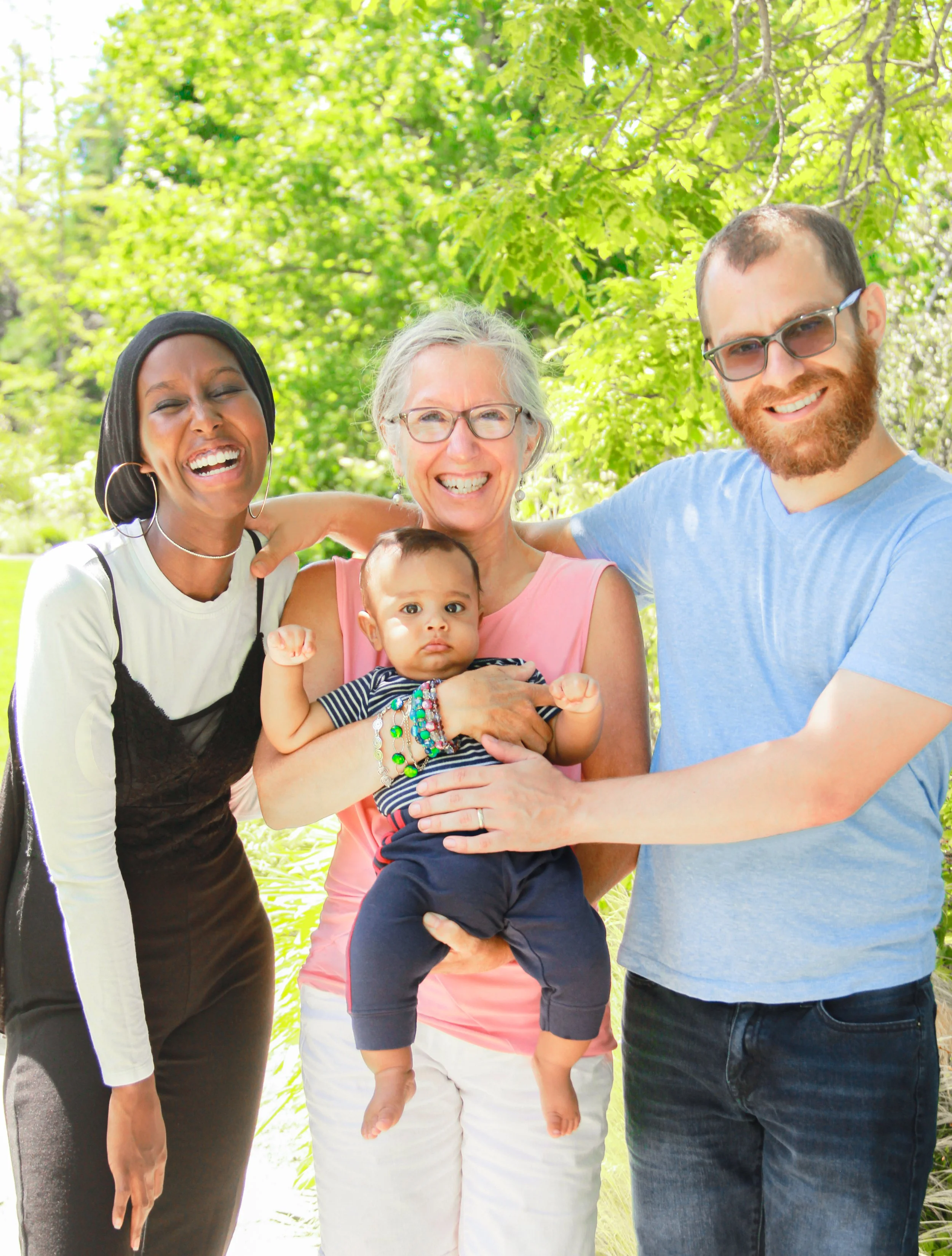 Group of four smiling adults, two women and two men, standing outdoors with green trees, holding a small child between them.
