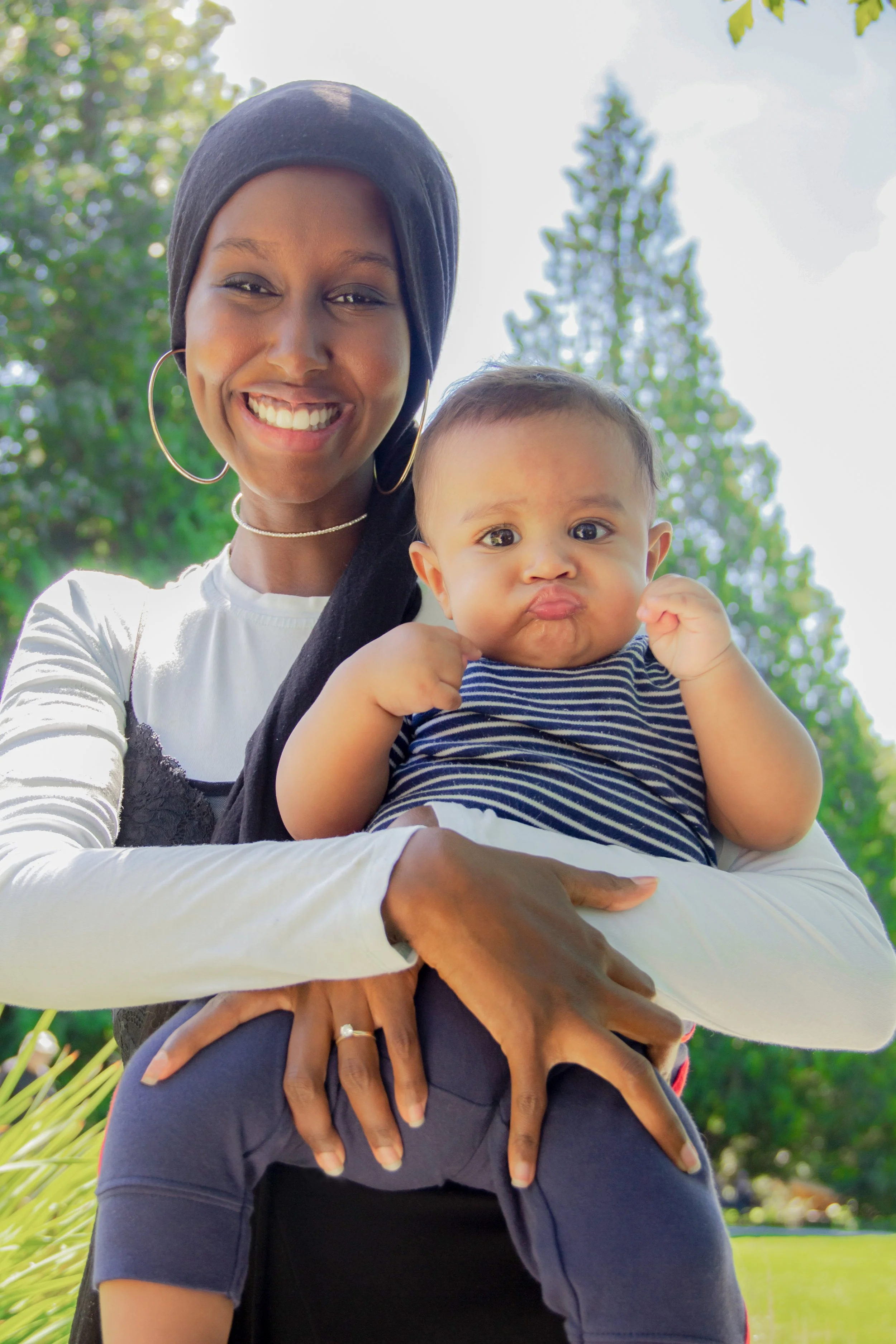 A smiling person holding a baby in a park setting with trees in the background. The person is wearing a black headscarf and hoop earrings, while the baby is wearing a striped shirt.