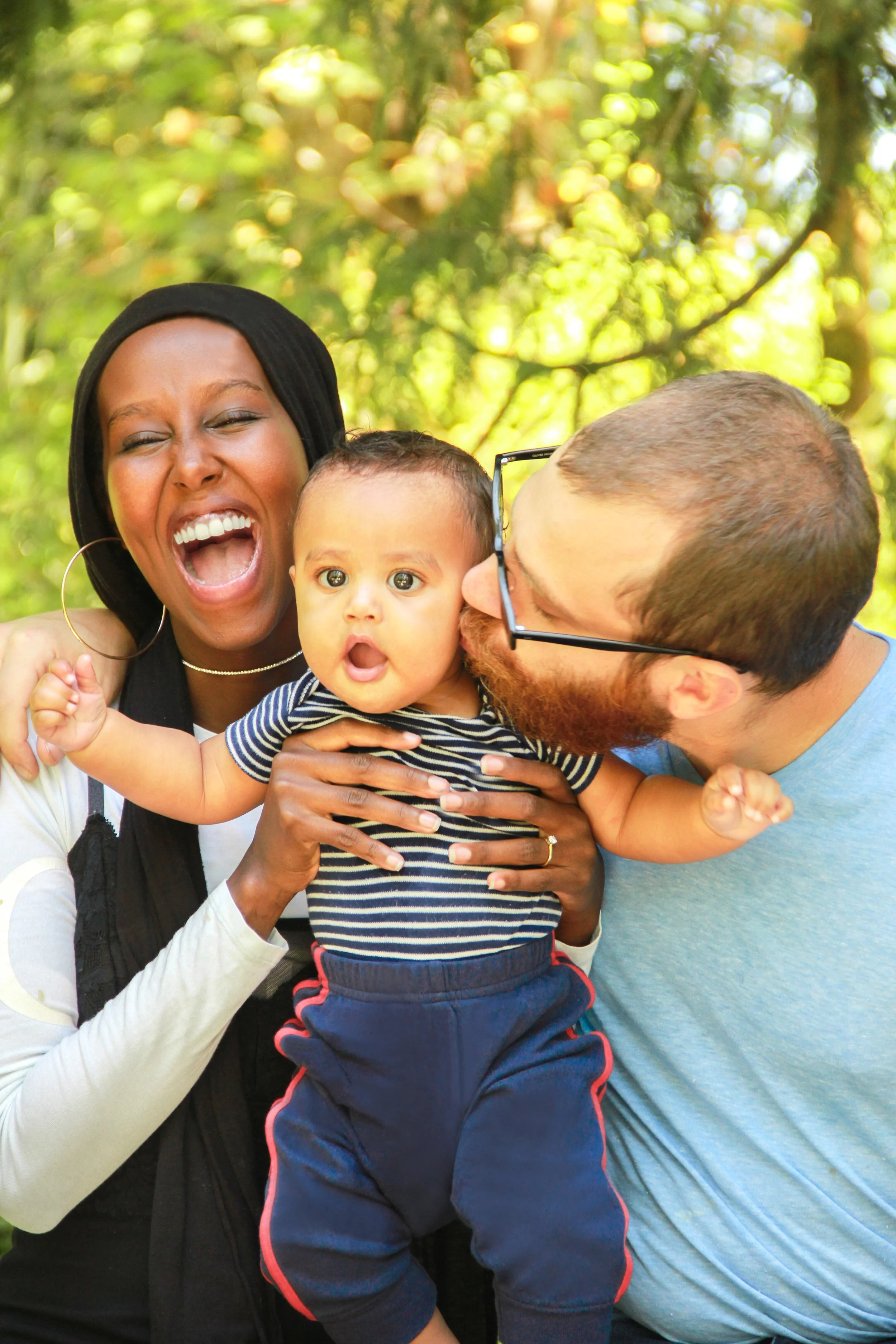 A joyful African-American woman and a Caucasian man kiss their baby boy in a park with green trees in the background.