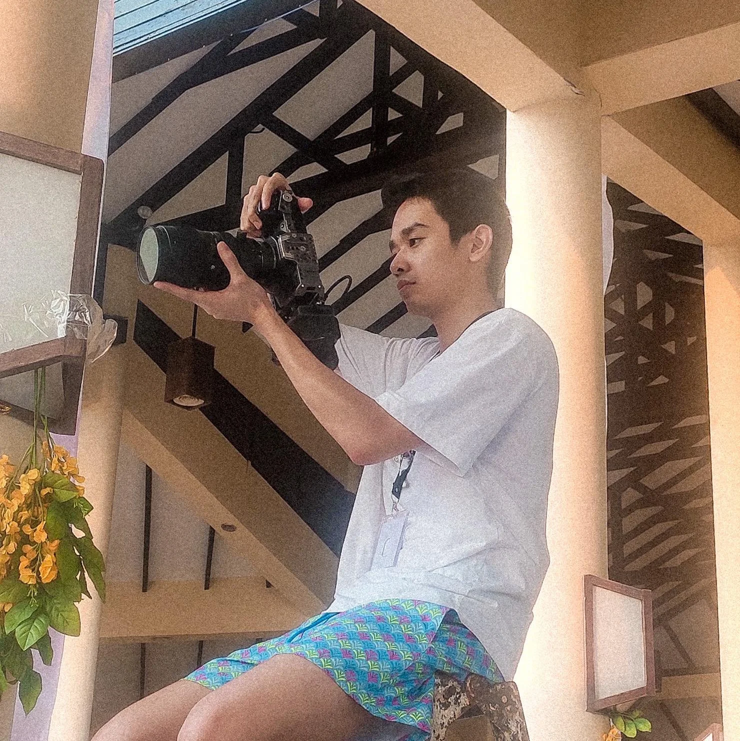 Person taking a photograph inside a building, wearing a white shirt and patterned shorts, holding a camera. Yellow flowers hanging in the foreground.