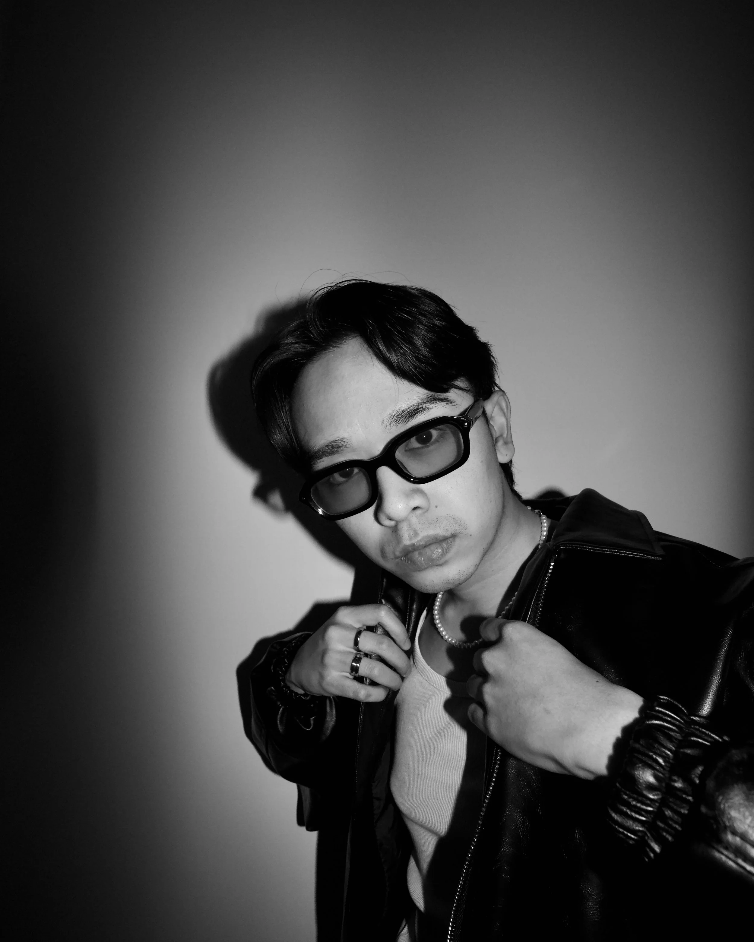 Black and white photo of a person wearing sunglasses and a leather jacket, adjusting the collar. The person is also wearing rings and a necklace.