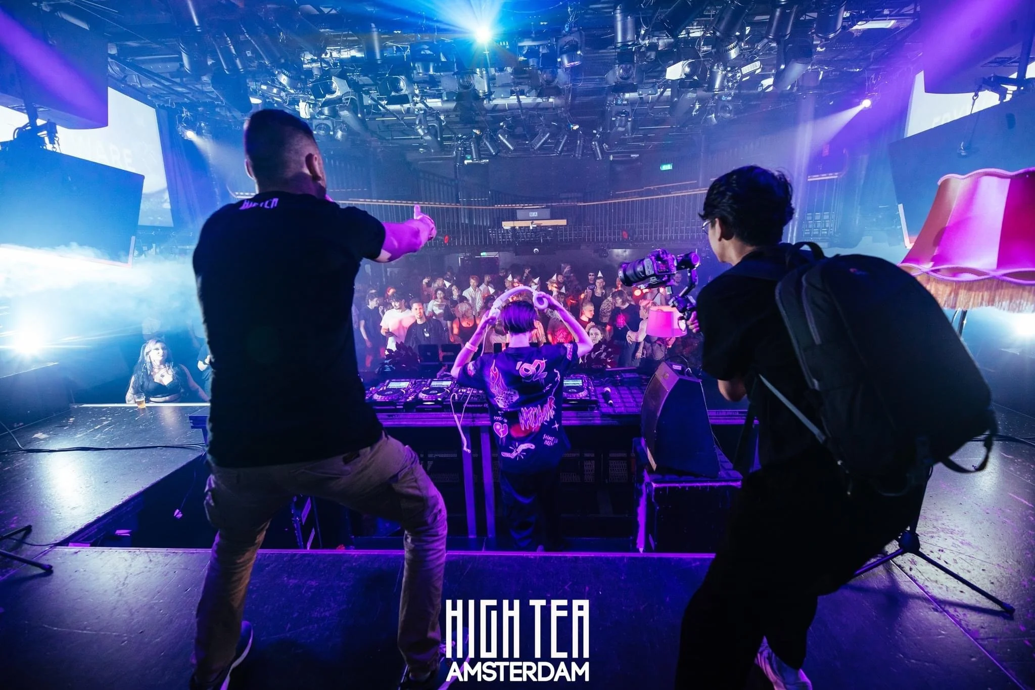 DJ performing on stage with a crowd at a club, photographer taking pictures, "High Tea Amsterdam" text visible.