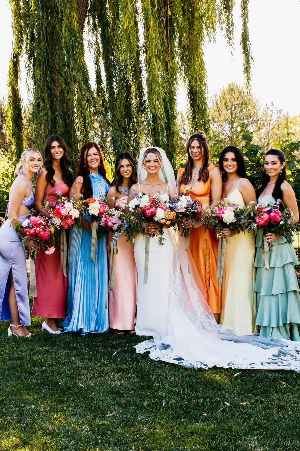 A bride in a white wedding dress and veil holding a bouquet of flowers, surrounded by eight bridesmaids in colorful dresses, all holding bouquets, standing outdoors on grass with green trees and foliage in the background.