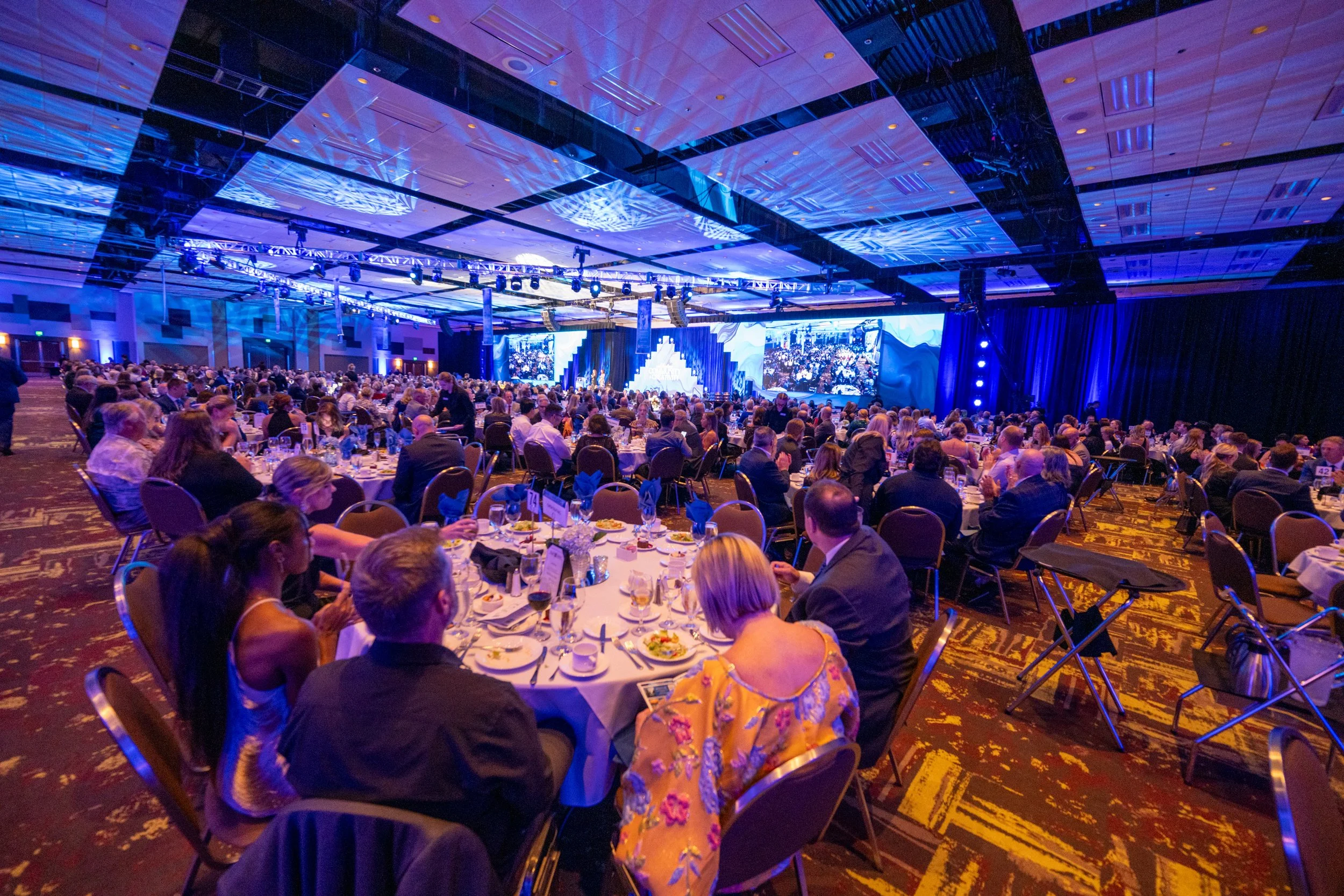 A large banquet hall filled with round tables of people, a stage with large screens, blue lighting, and ceiling decorations, hosting a formal event or conference.