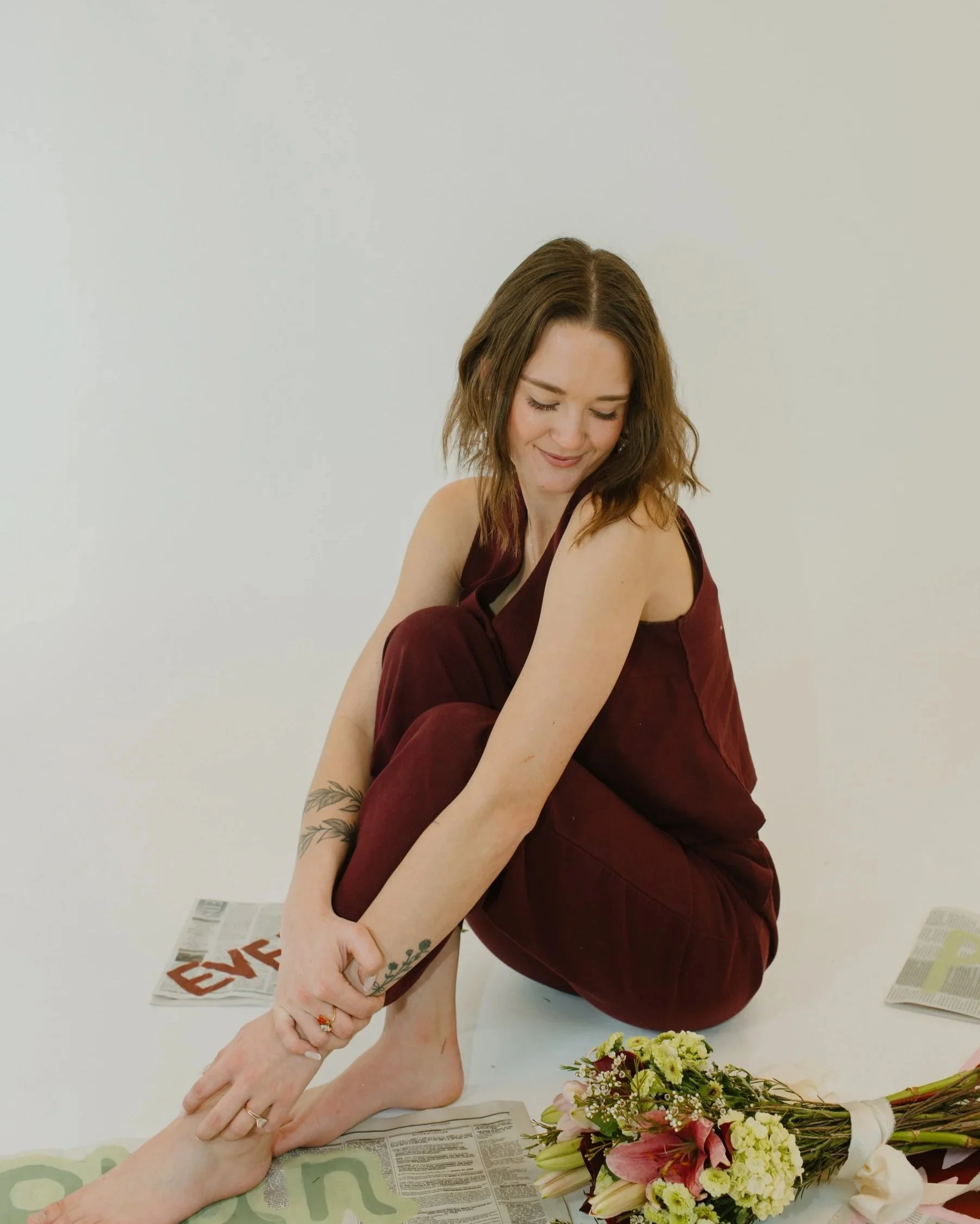 A woman with brown hair and tattoos on her arm, smiling while sitting on the floor. She is dressed in maroon overalls, holding her foot, with newspapers and a bouquet of flowers nearby.