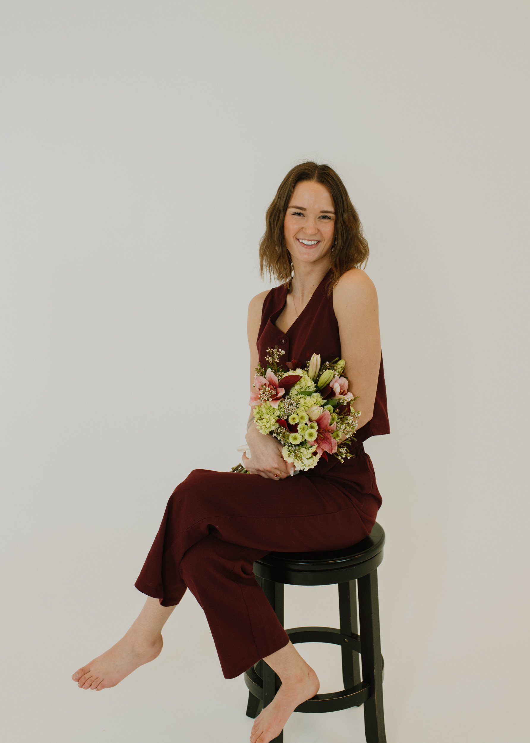 A woman sitting on a black stool, holding a bouquet of flowers, smiling, against a plain white background.