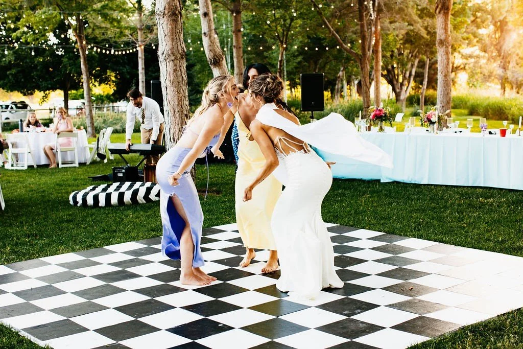 A bride and two women dancing on a black and white checkered dance floor outdoors during a wedding reception, with guests sitting at tables and string lights hanging overhead.