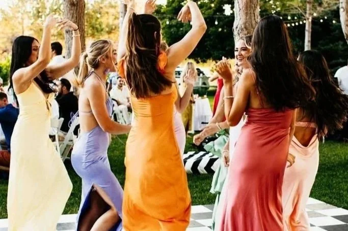 Group of women in colorful dresses dancing and having fun outdoors at a garden party with string lights.