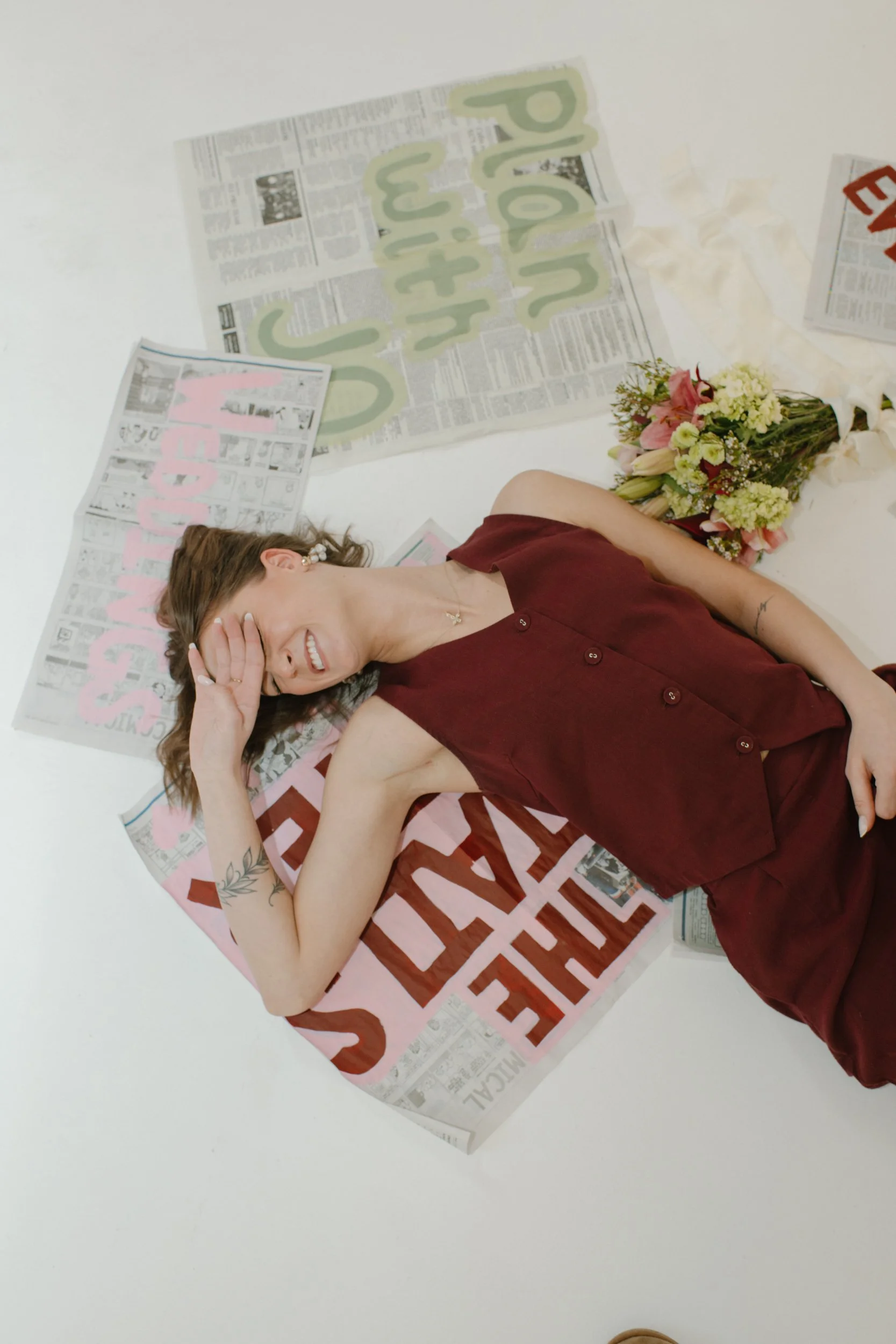A woman lying on the floor, smiling with her hand covering her face, surrounded by flowers, newspapers, and colorful signs.