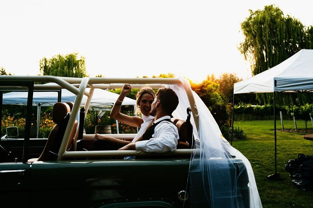 A newlywed couple sitting in a vintage car with a veil, celebrating outdoors at sunset.