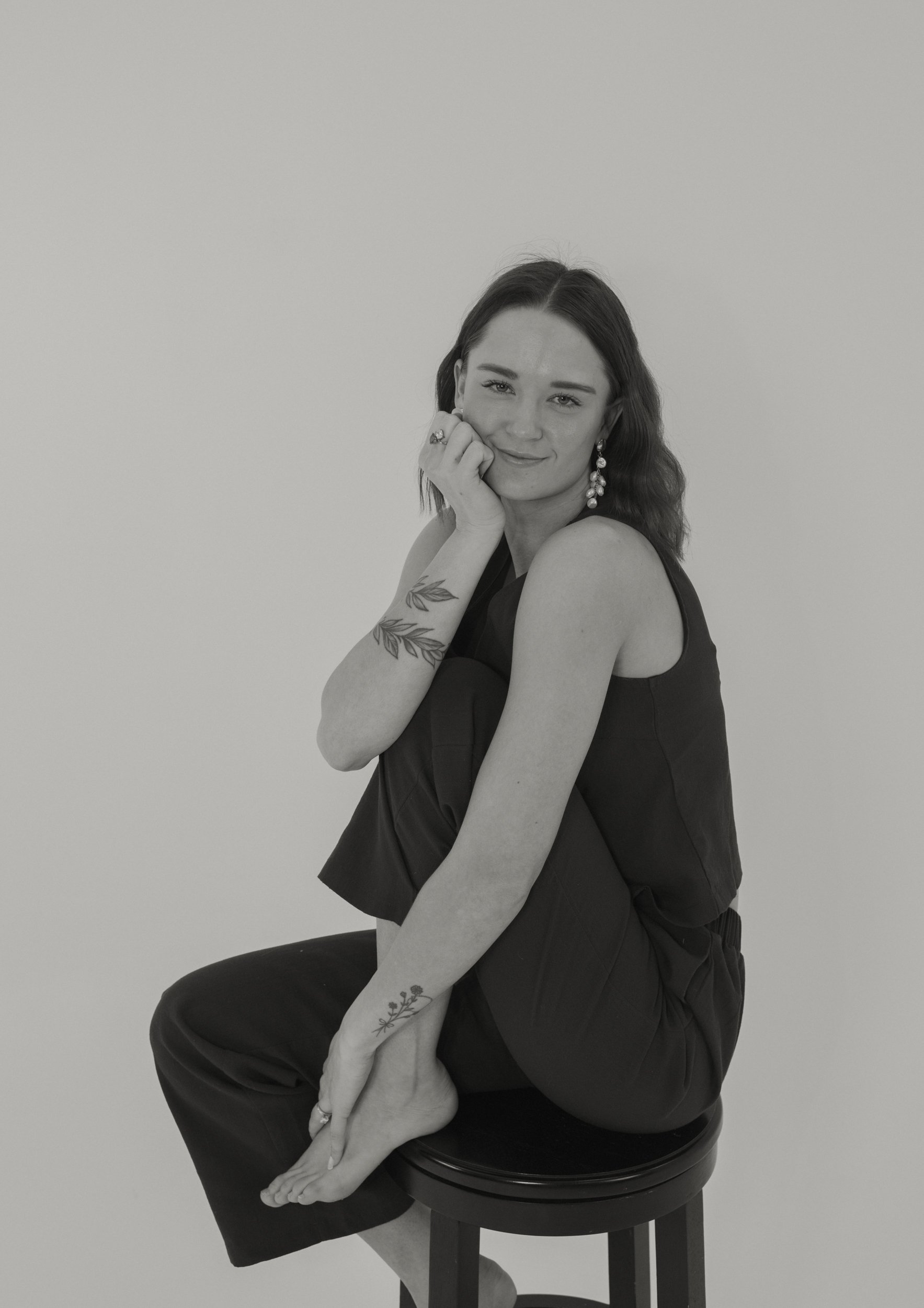 Black and white portrait of a woman sitting on a stool with one leg crossed over the other, smiling gently, with her hand resting on her face.
