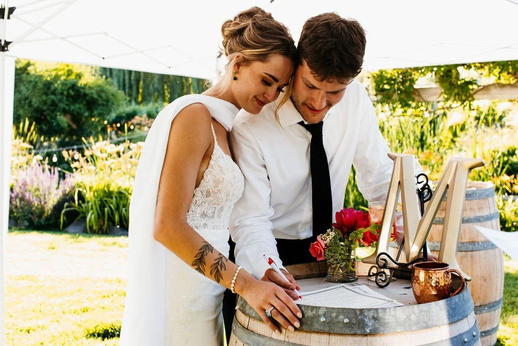 Bride and groom signing marriage papers outdoors under a white canopy with flowers and decorations on a wooden barrel table.