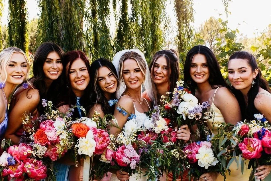 Group of women smiling and holding bouquets of flowers outdoors, with trees and greenery in the background.
