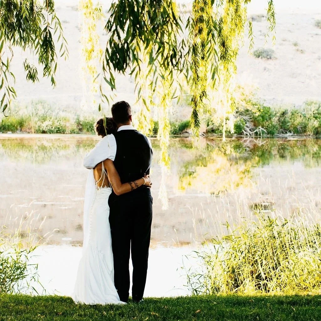 A couple, dressed in wedding attire, embraces beside a lake under a tree with hanging branches.