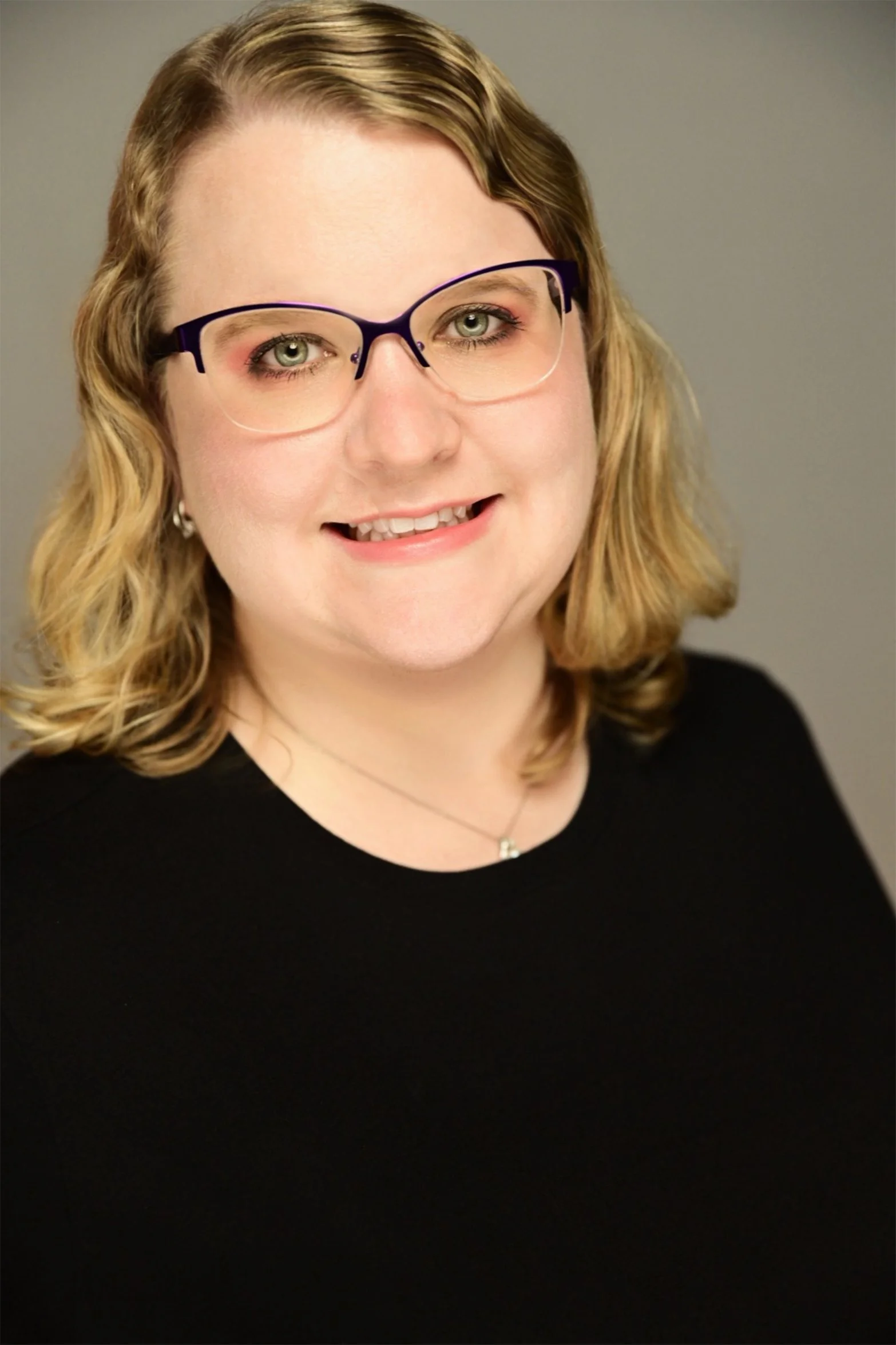 A woman with blonde hair, green eyes, smiles at the camera. She is wearing purple glasses and a black t-shirt.