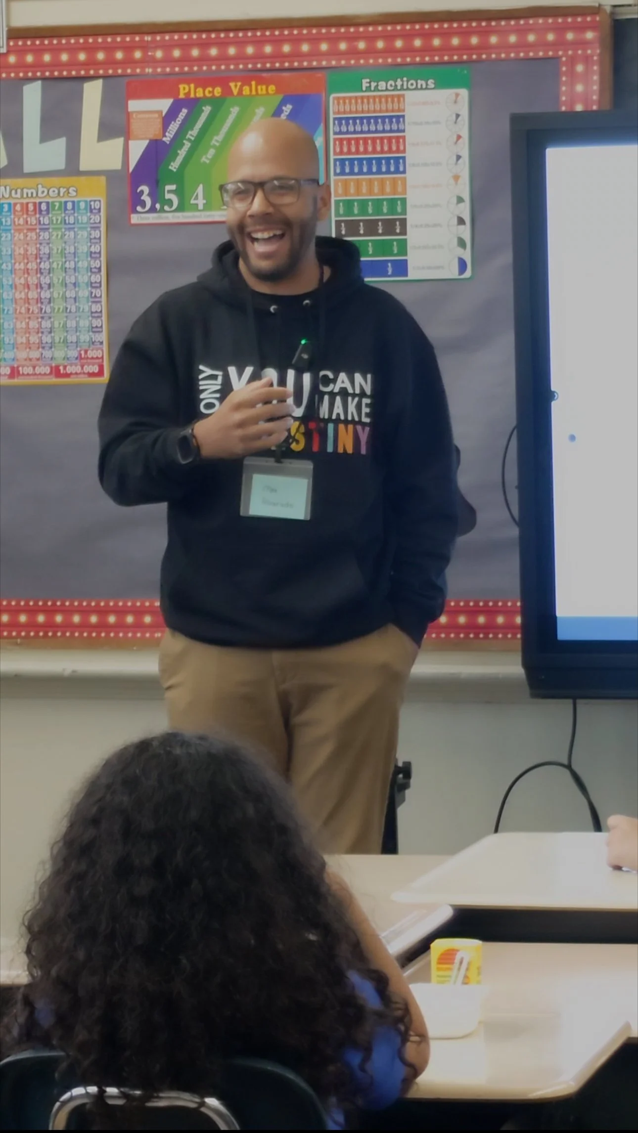 A teacher in a black hoodie with colorful text stands at the front of a classroom, smiling and holding a microphone. Behind him are educational posters about place value and fractions, and a large digital screen. A student with curly hair is seated at a desk in the foreground.