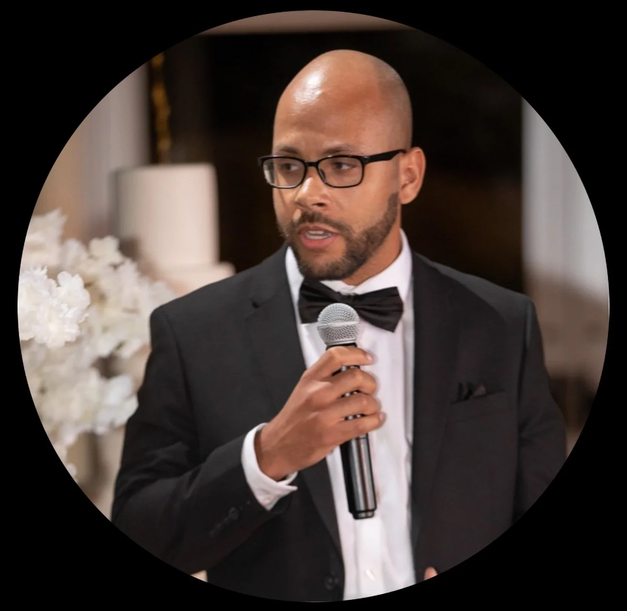 Man in a tuxedo and glasses speaking into a microphone at a formal event, with white flowers and a blurred background.
