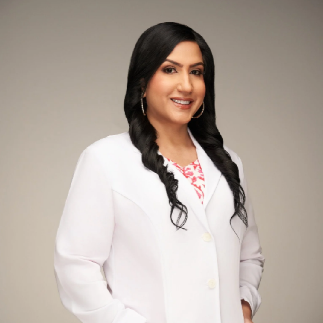 Portrait of a smiling woman with long black hair, wearing hoops, and a white blazer over a pink patterned top, standing against a neutral background.