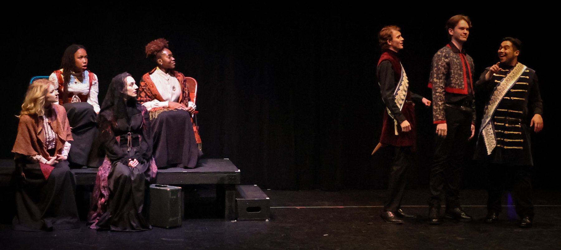 Theatrical performance with actors in period costumes on stage, some sitting and some standing, in a dramatic scene.