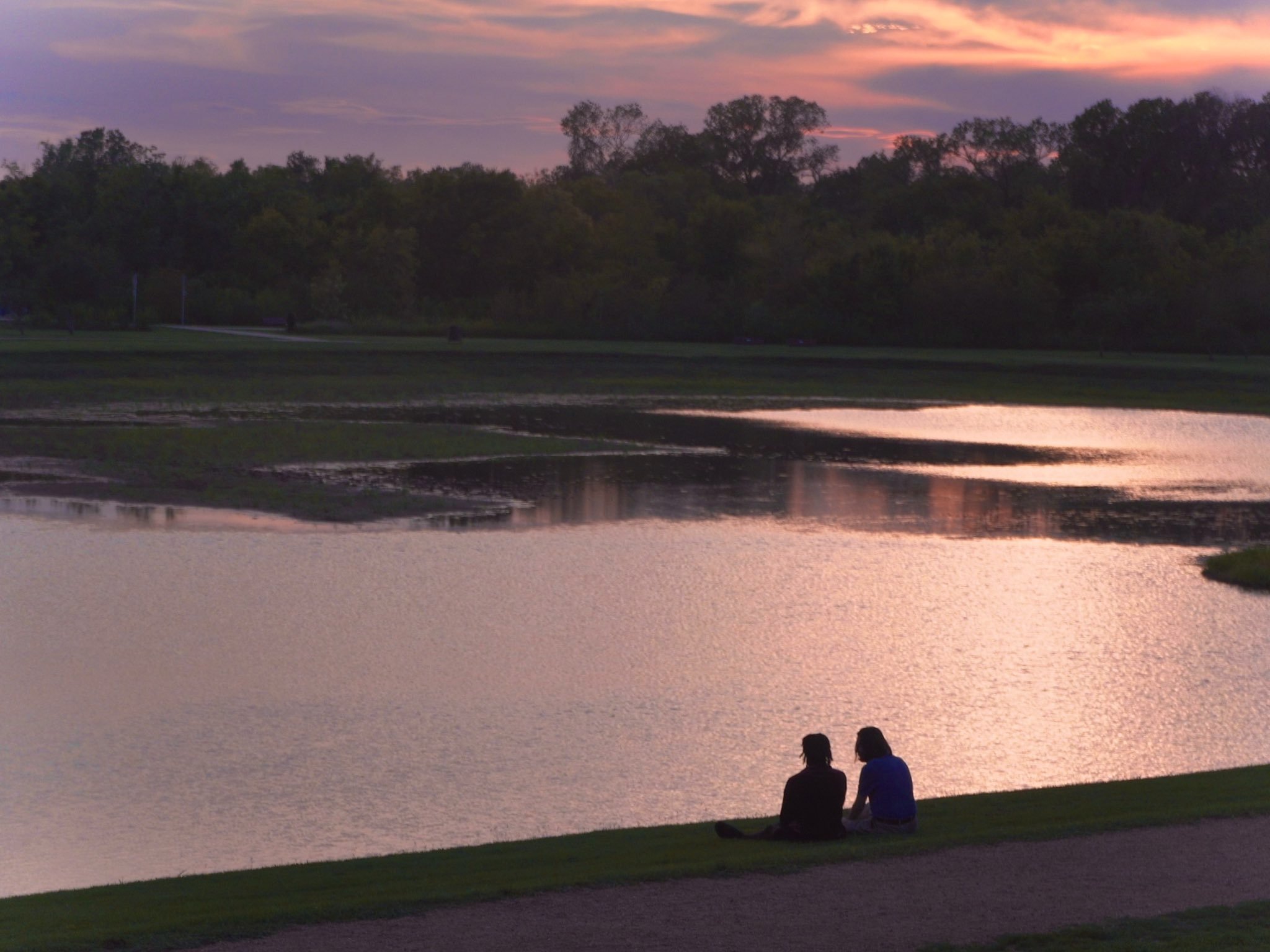 Two people sitting by a lake at sunset, with trees and a colorful sky in the background.