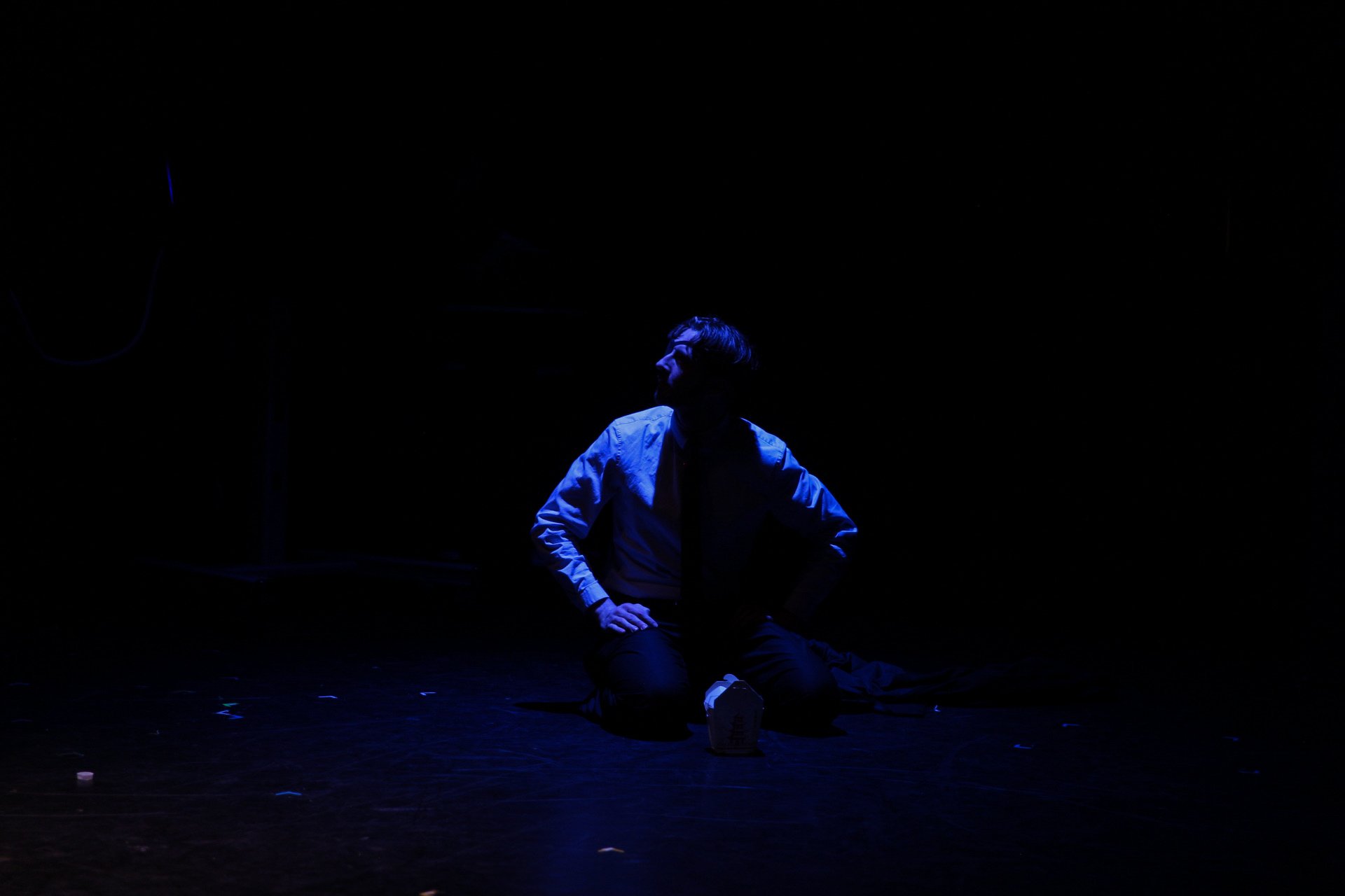 Person kneeling on a stage in dim blue light, wearing a shirt and tie, with dark surroundings.