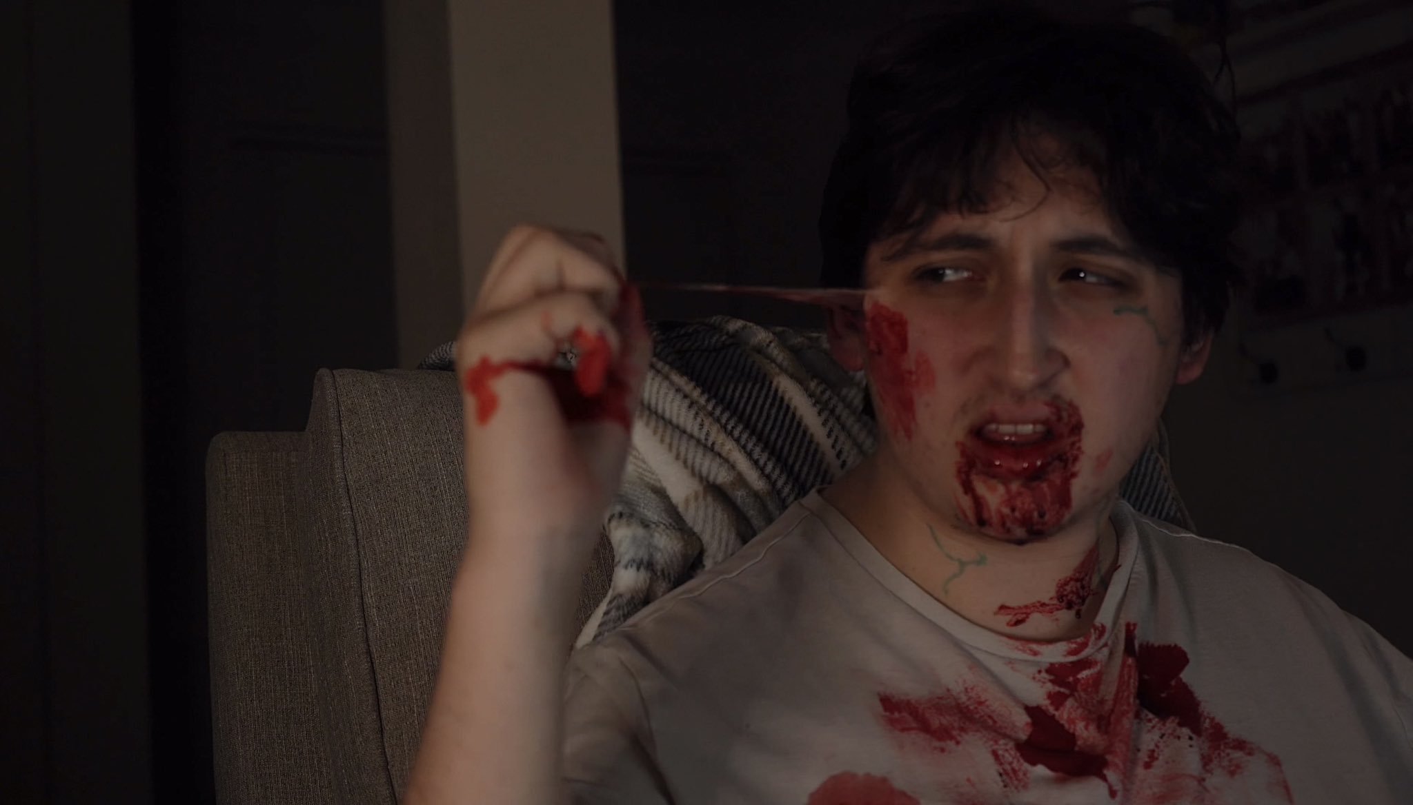 Person sitting on a chair with fake blood on their face and shirt, holding a bloodied object.