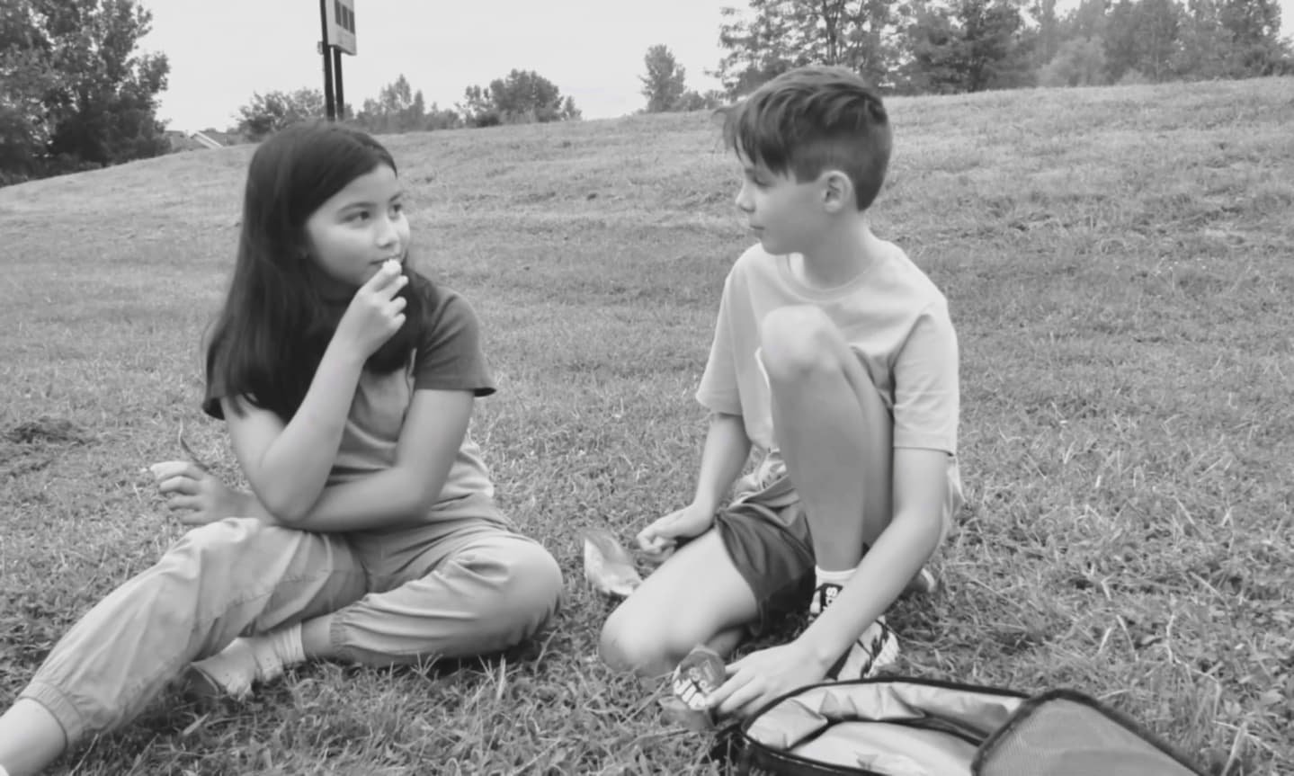 Two kids sitting on grass, talking, with a backpack and snack packet beside them.