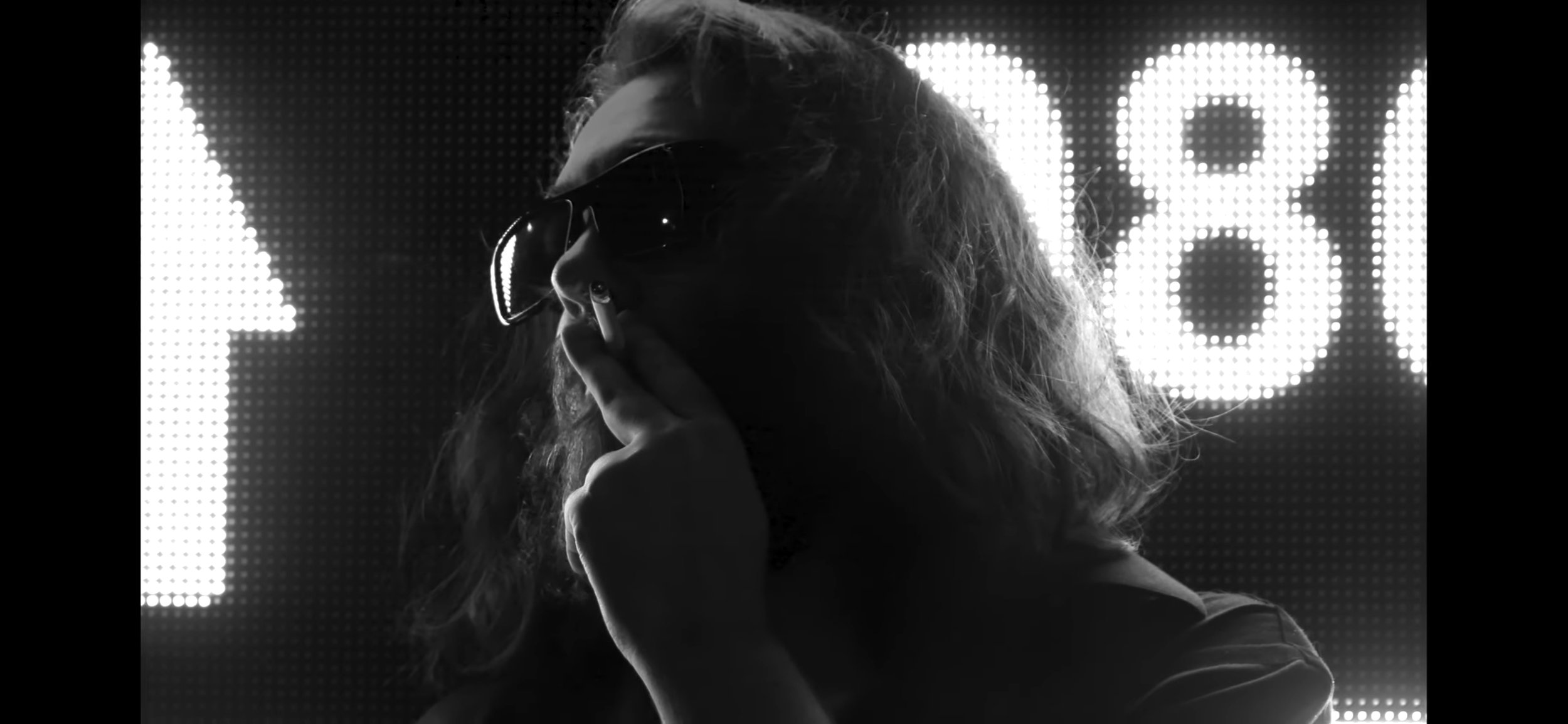 Black and white photo of a person with long hair wearing sunglasses, smoking a cigarette in front of an LED screen displaying an arrow and number 808.
