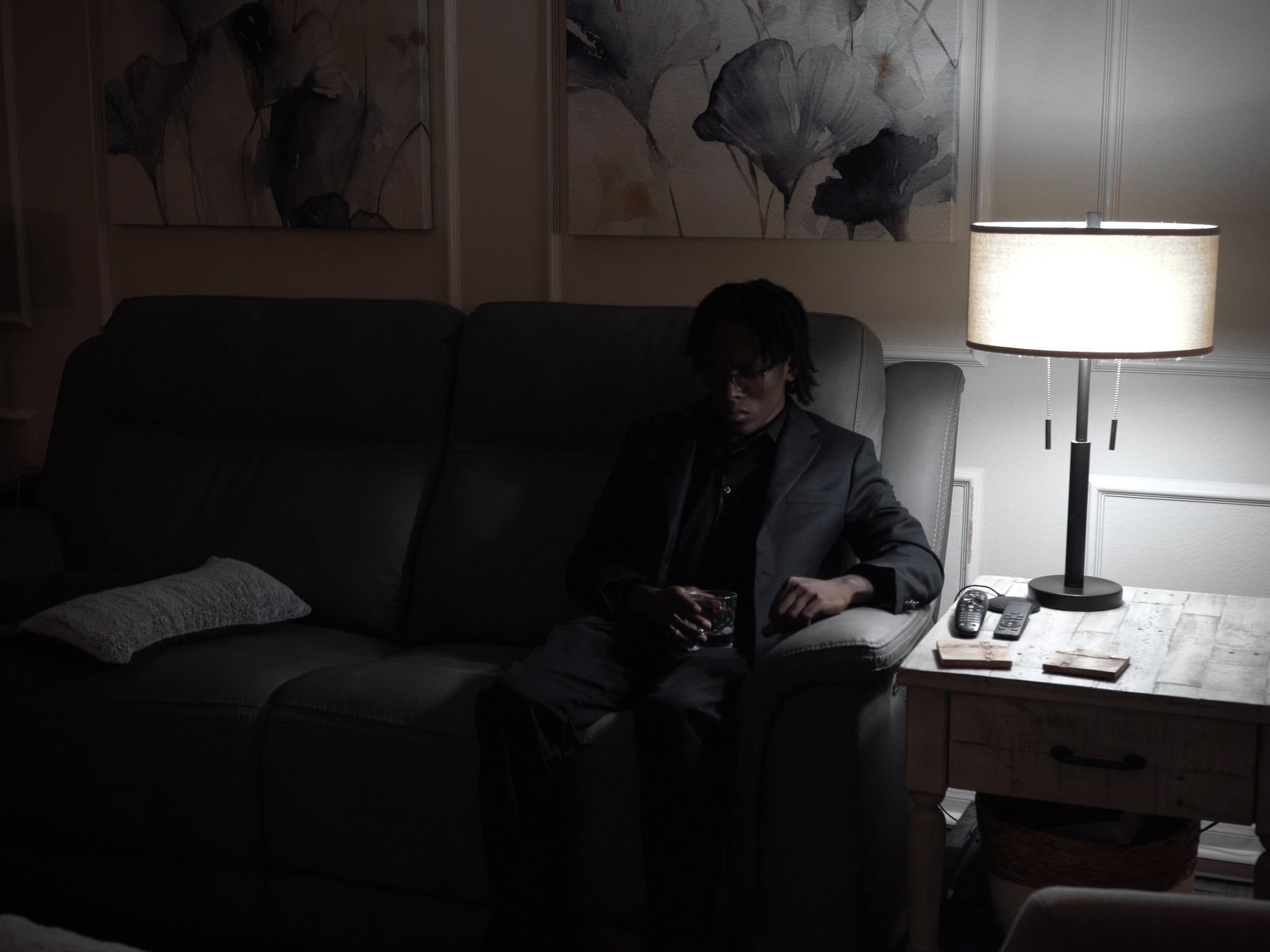 Person in a dark room sitting on a sofa near a side table with a lamp and remote controls.