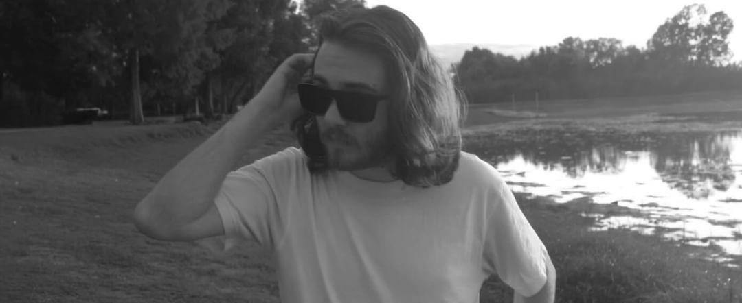 Man with long hair and sunglasses standing near water in a park, black and white photo.