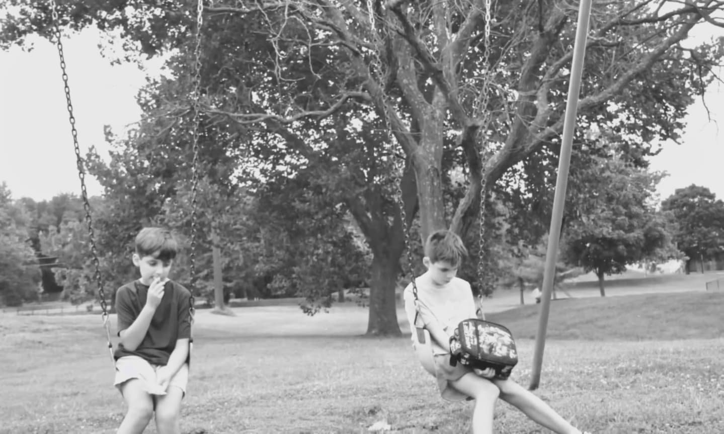 Two children sitting on swings in a park with trees and grass in the background.