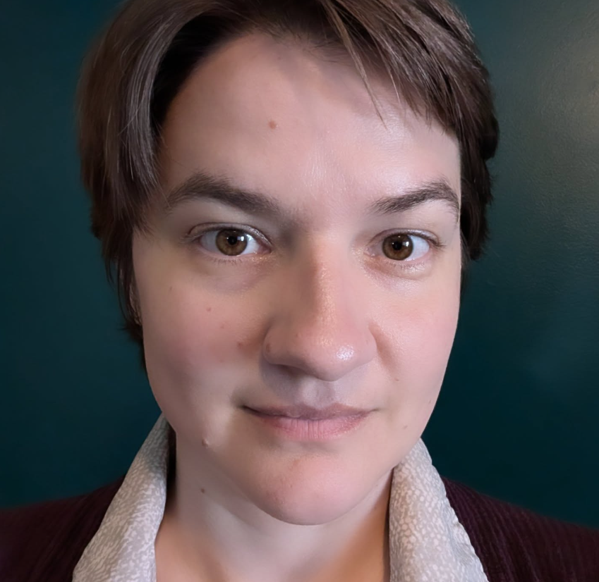 Close-up of a person with short brown hair, light skin, and brown eyes, wearing a collared shirt and a dark jacket, standing against a teal background.