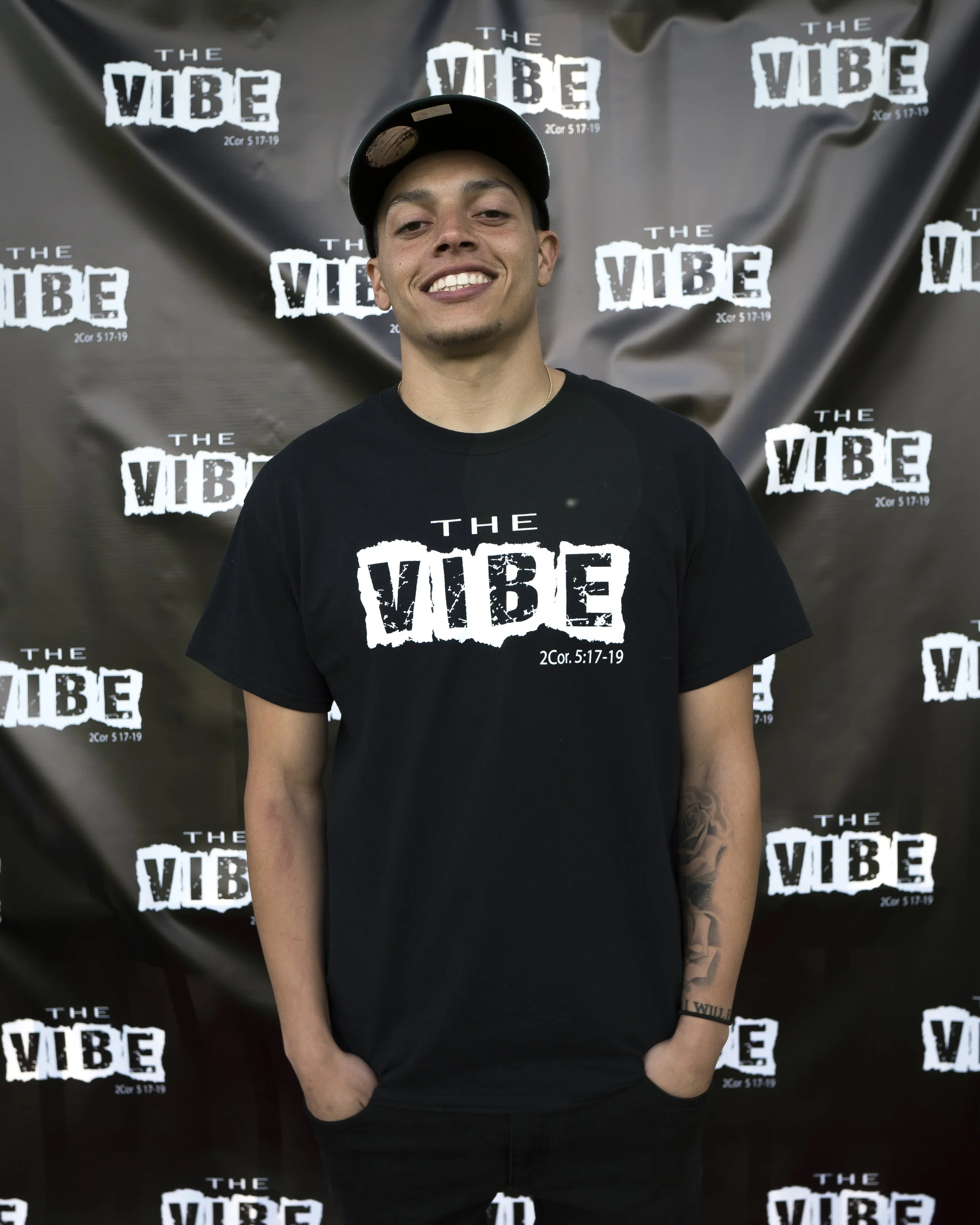Young man smiling, wearing a black T-shirt and cap, standing in front of a backdrop with multiple 'The VIBE' logos and a Bible verse reference.