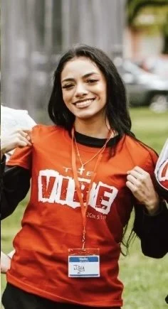 Young woman with dark hair, smiling, wearing a red Volunteer t-shirt and a name badge, standing outdoors on grass with buildings and cars in the background.