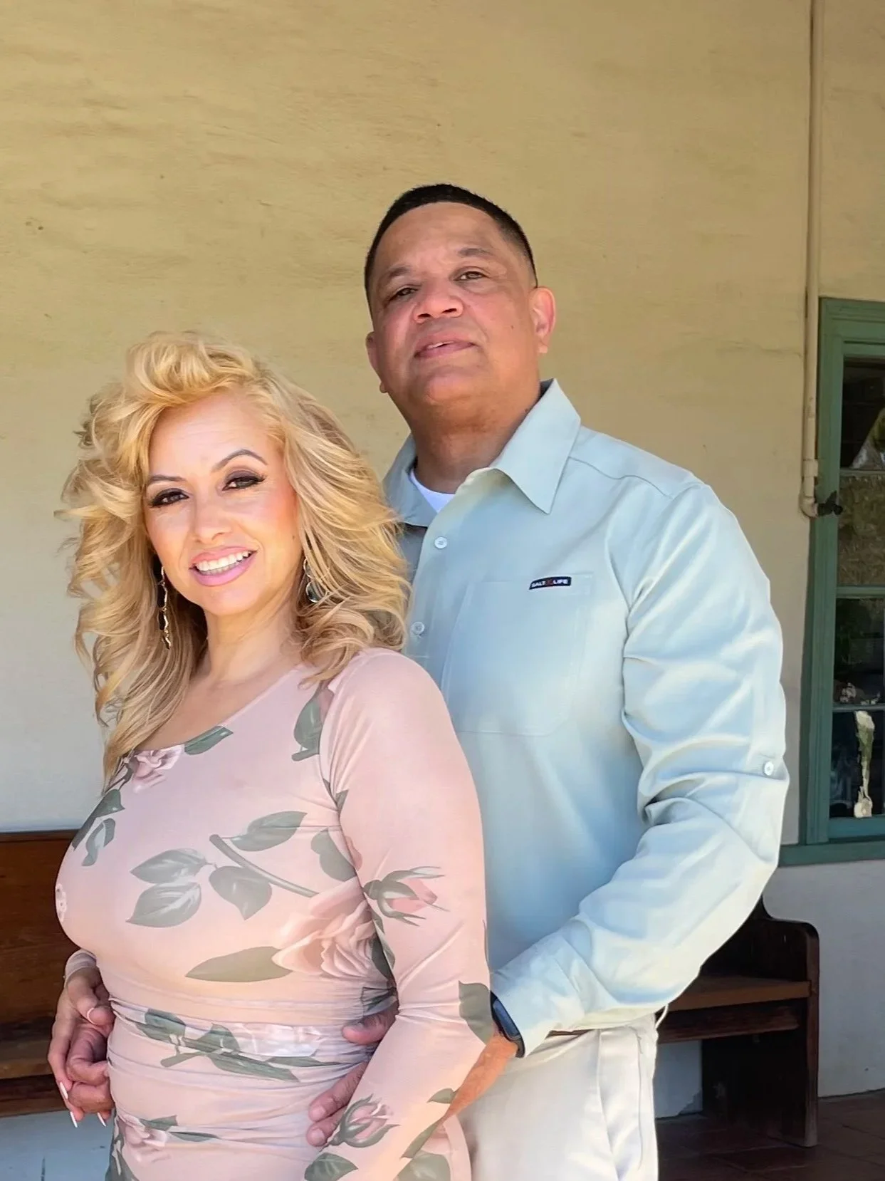 A woman with blonde, curly hair wearing a pink floral dress standing next to a man with short dark hair wearing a light-colored button-up shirt.