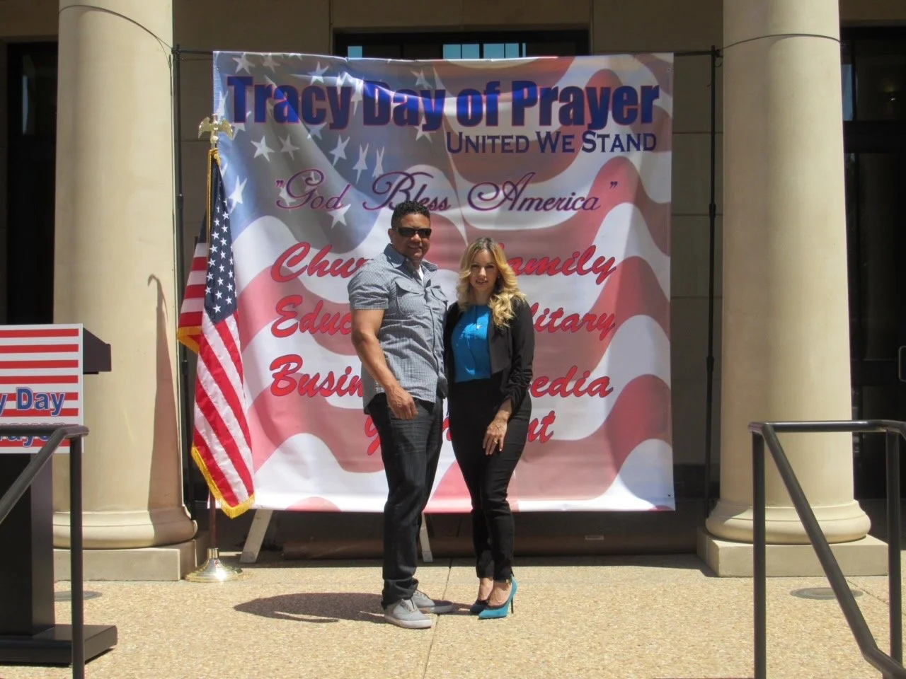 Two people standing in front of a patriotic banner for 'Tracy Day of Prayer' with an American flag beside them, on a sunny day outside a building.