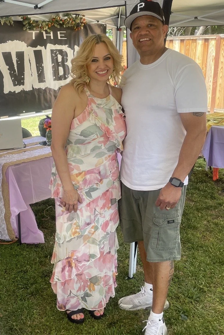 A woman with blonde hair wearing a floral dress and a man wearing a white t-shirt, shorts, and a Pittsburgh Pirates cap, standing together outdoors under a canopy, smiling for the camera.