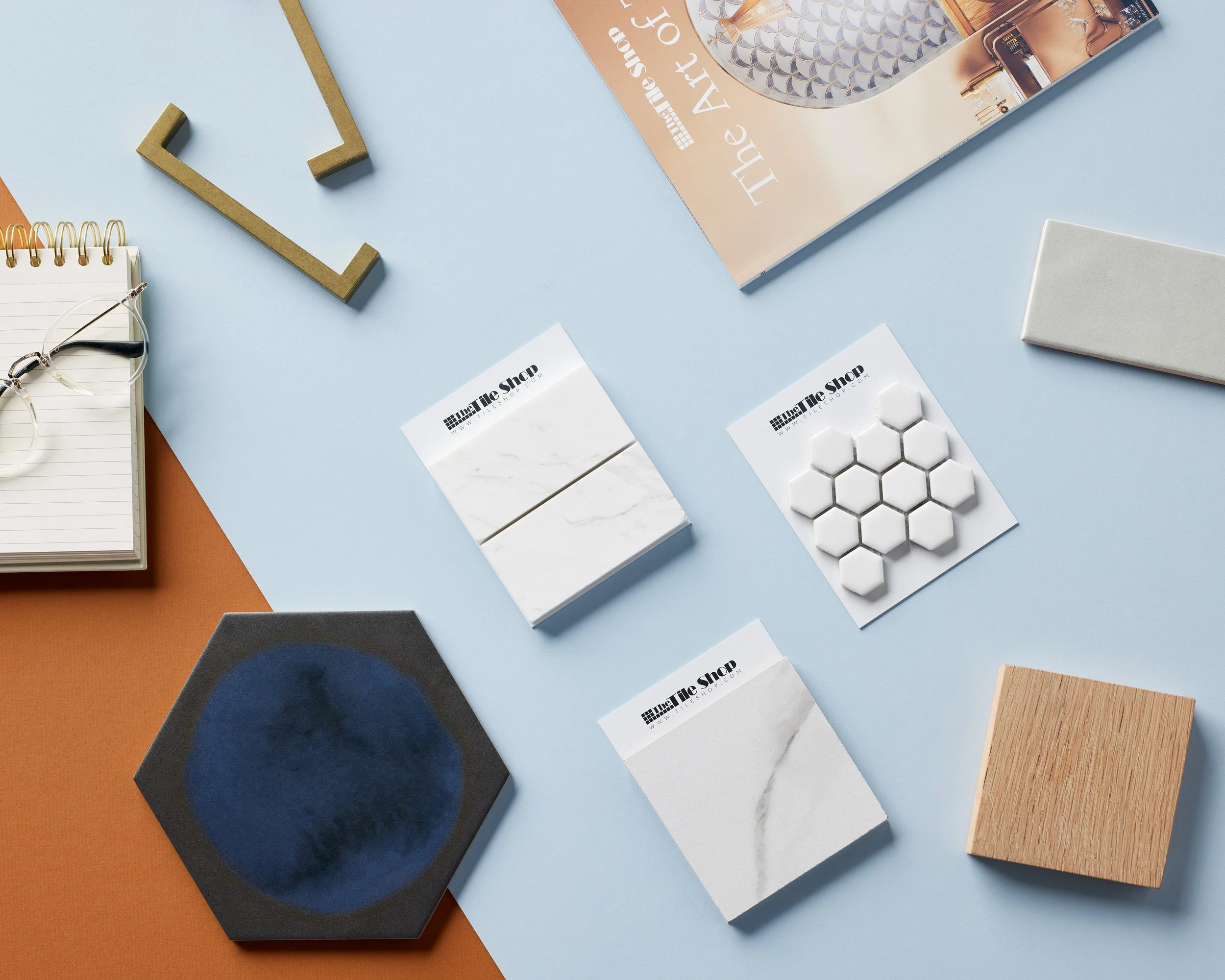 A flat lay of various home decor and design samples, including tiles, a hexagonal blue tray, a small stack of magazines and a notebook with glasses on top, all arranged on a table with blue and orange sections.