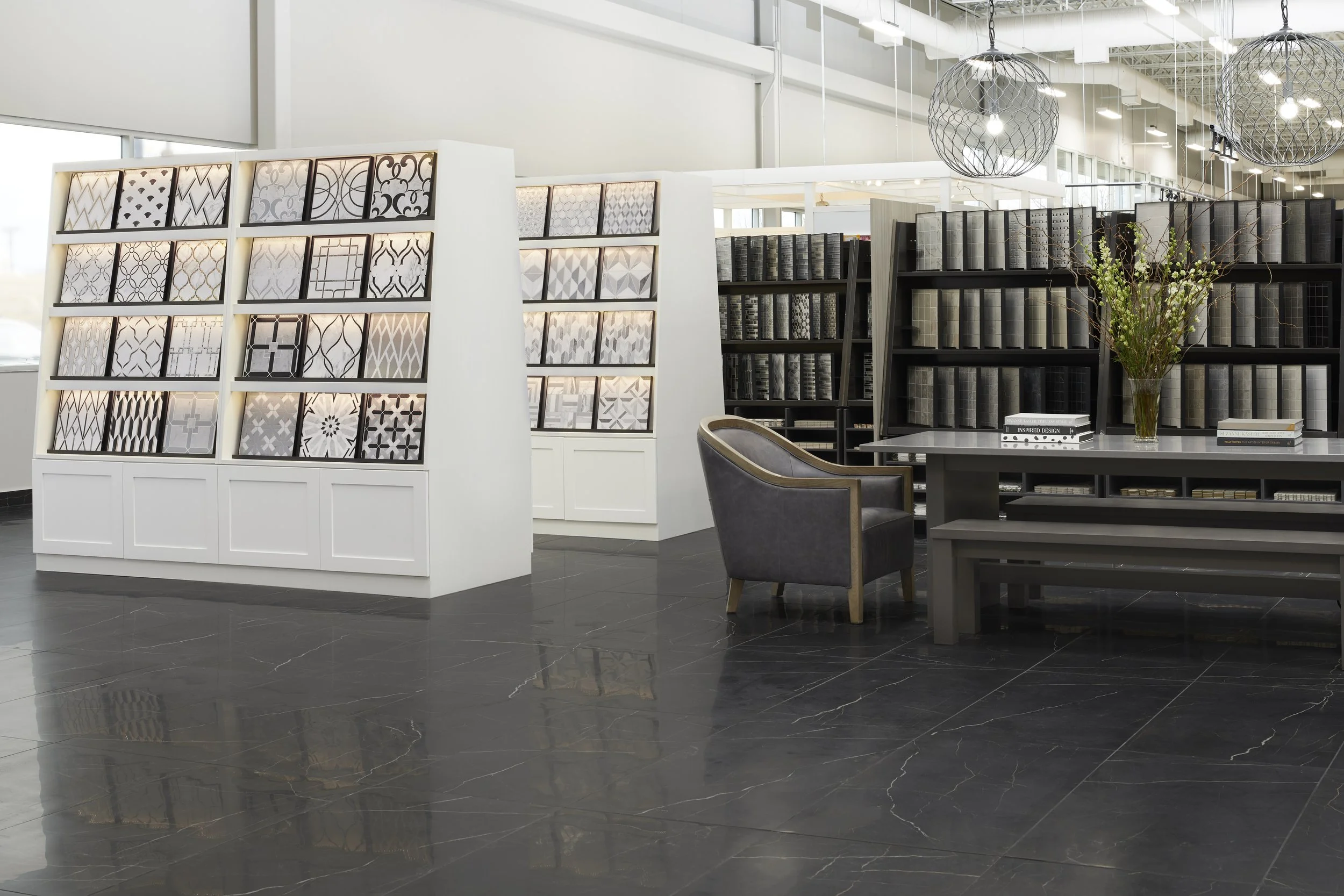 Interior of a modern showroom displaying various tile samples on white and black shelves, with a black armchair, a table with books, and decorative pendant lights hanging from the ceiling.