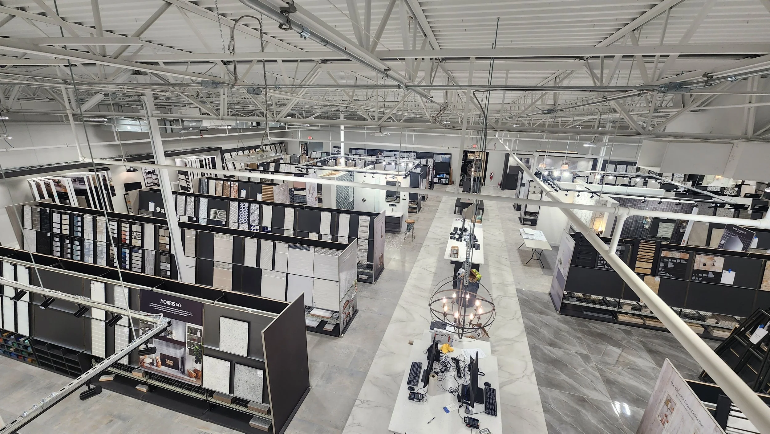 View of a large showroom displaying various tile samples and materials, with computer workstations and industrial lighting fixtures overhead.