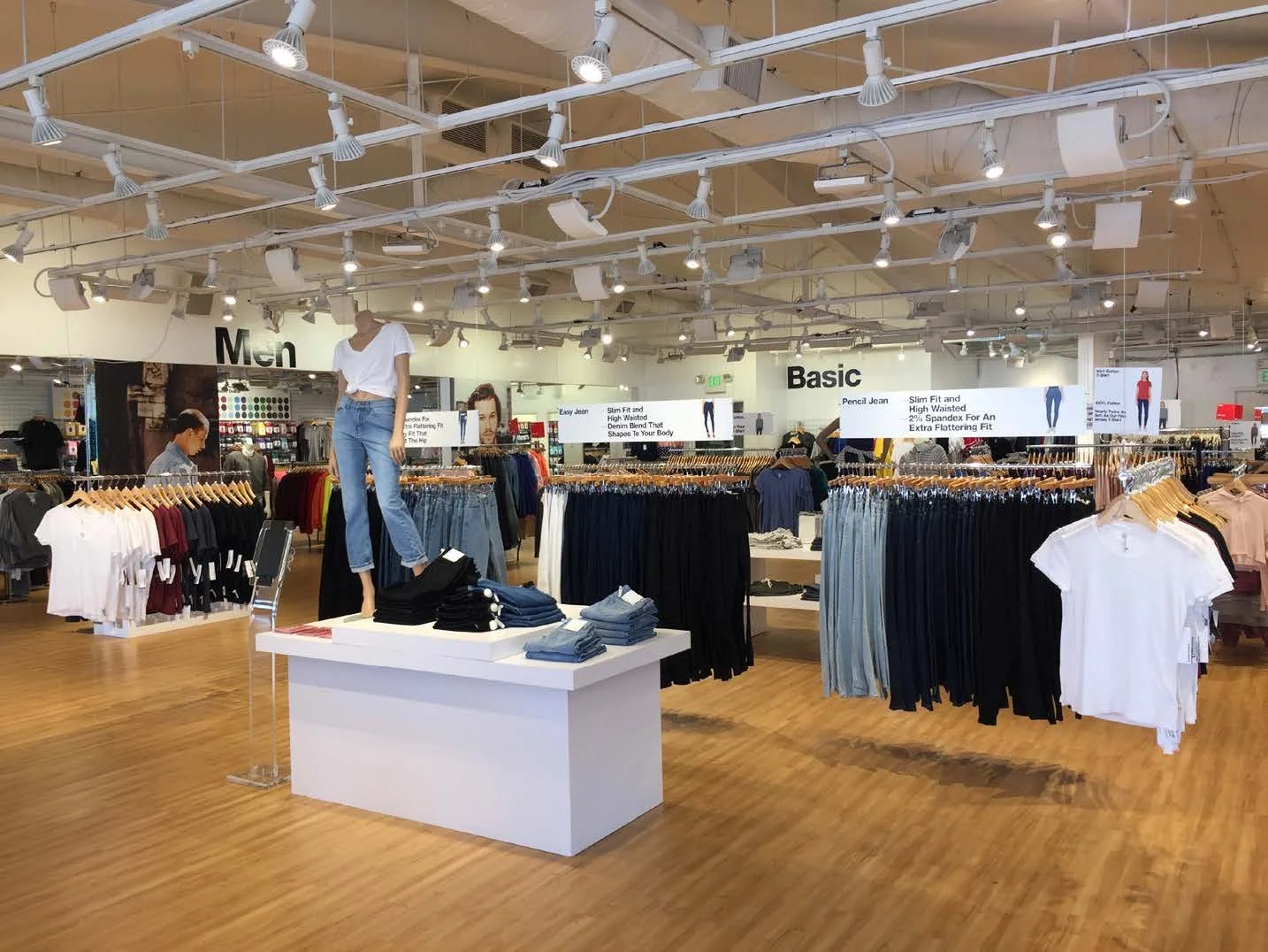 Clothing store with tables and racks of jeans and t-shirts, mannequin wearing jeans and a white shirt, signs indicating 'Men' and 'Basic' sections, and overhead lighting