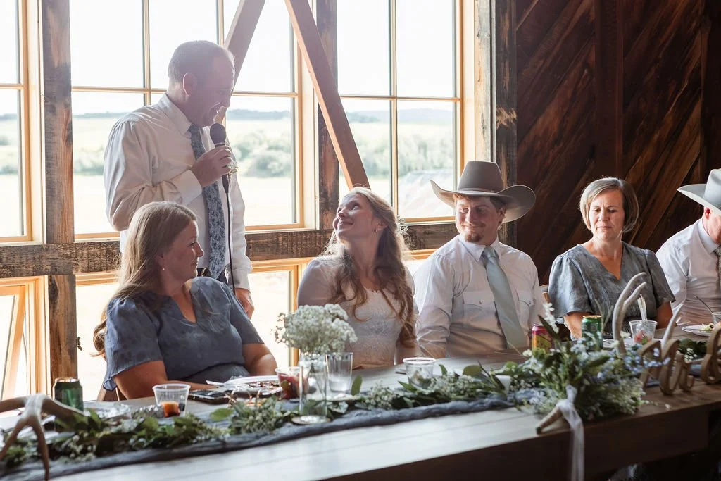 A man giving a speech at a wedding reception with guests seated at a decorated table, including a woman in a white dress and several people wearing cowboy hats.