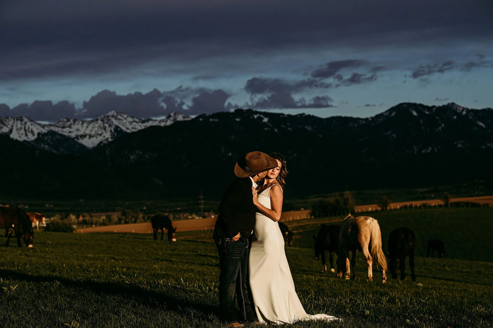 A couple stands close together in a grassy field during sunset, with mountains and dark clouds in the background. The woman wears a white dress, and the man wears a cowboy hat and dark clothing.