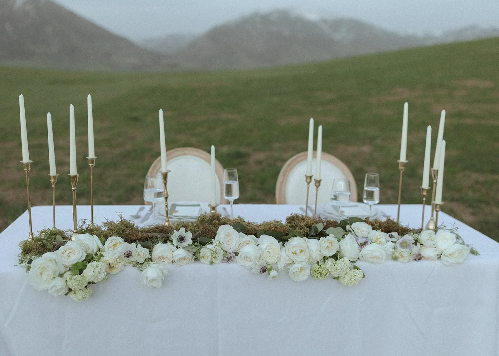 Elegant outdoor wedding reception table decorated with white roses, candles, and greenery, set against a grassy hillside with mountains in the background.